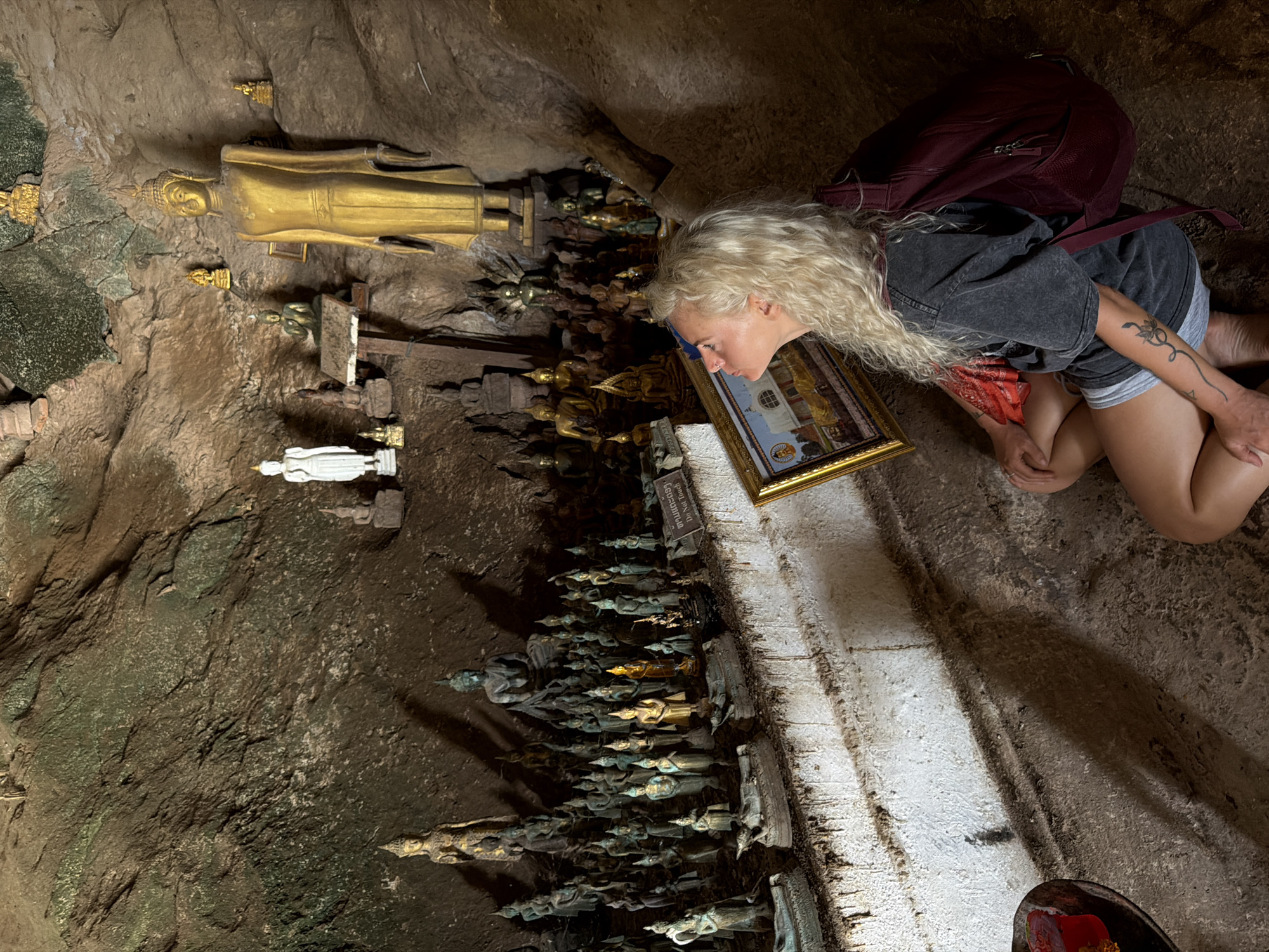 In the city of Muangkeo, Laos, within a cave, a woman with long, curly blonde hair sits on the cave floor, looking upwards. She is wearing a dark grey t-shirt, grey shorts, and a maroon backpack. In front of her, she holds a framed painting with a golden border. The cave walls are rough and textured. Throughout the cave, there are numerous small Buddha statues and several larger golden and white Buddha statues. One prominent golden Buddha statue stands tall on the right side of the image. The floor of the cave appears to be dirt and stone.