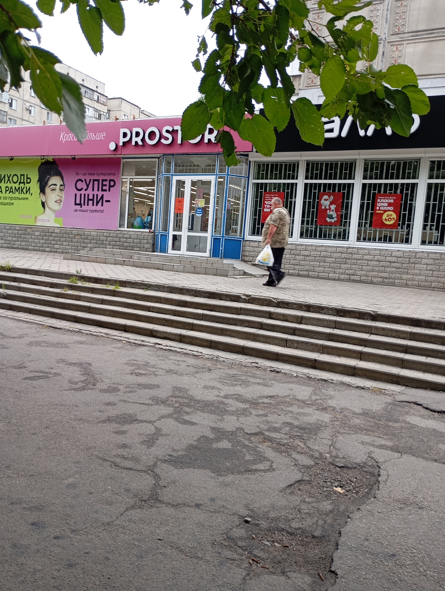 The image captures a street scene in Horishni Plavni, Ukraine. In the foreground, a cracked asphalt road leads towards a paved sidewalk and a set of concrete stairs. A man, wearing a camouflage-patterned shirt and dark pants, walks away from the viewer on the sidewalk, carrying a white plastic bag. Behind him is a pink and white building with the store name "PROSTOR" prominently displayed. The store features large windows, some of which display posters, including one with a red background and the text "до -60%". The building appears to be a commercial establishment, possibly a supermarket or department store, with several other windows and an entrance. Overhanging the scene are branches with green leaves, obscuring parts of the sky and the upper portions of the buildings. The overall environment suggests an urban setting on a cloudy day.