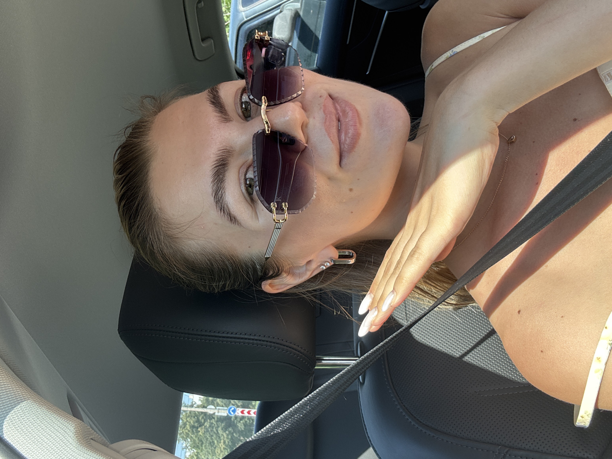 A young Caucasian woman with blonde hair pulled back, wearing large, dark-lensed sunglasses, and a light-colored, floral-patterned top, is pictured inside a car. Her right hand, with long white nails, is raised to her face, partially obscuring her chin and neck. She is wearing a silver hoop earring with two small stones. The interior of the car is visible, including a black leather seat and a seatbelt. Sunlight casts shadows on her skin and the seat. Outside the car, a street with trees and traffic signs can be seen through the windows. The woman appears to be looking towards the camera with a slight smile.