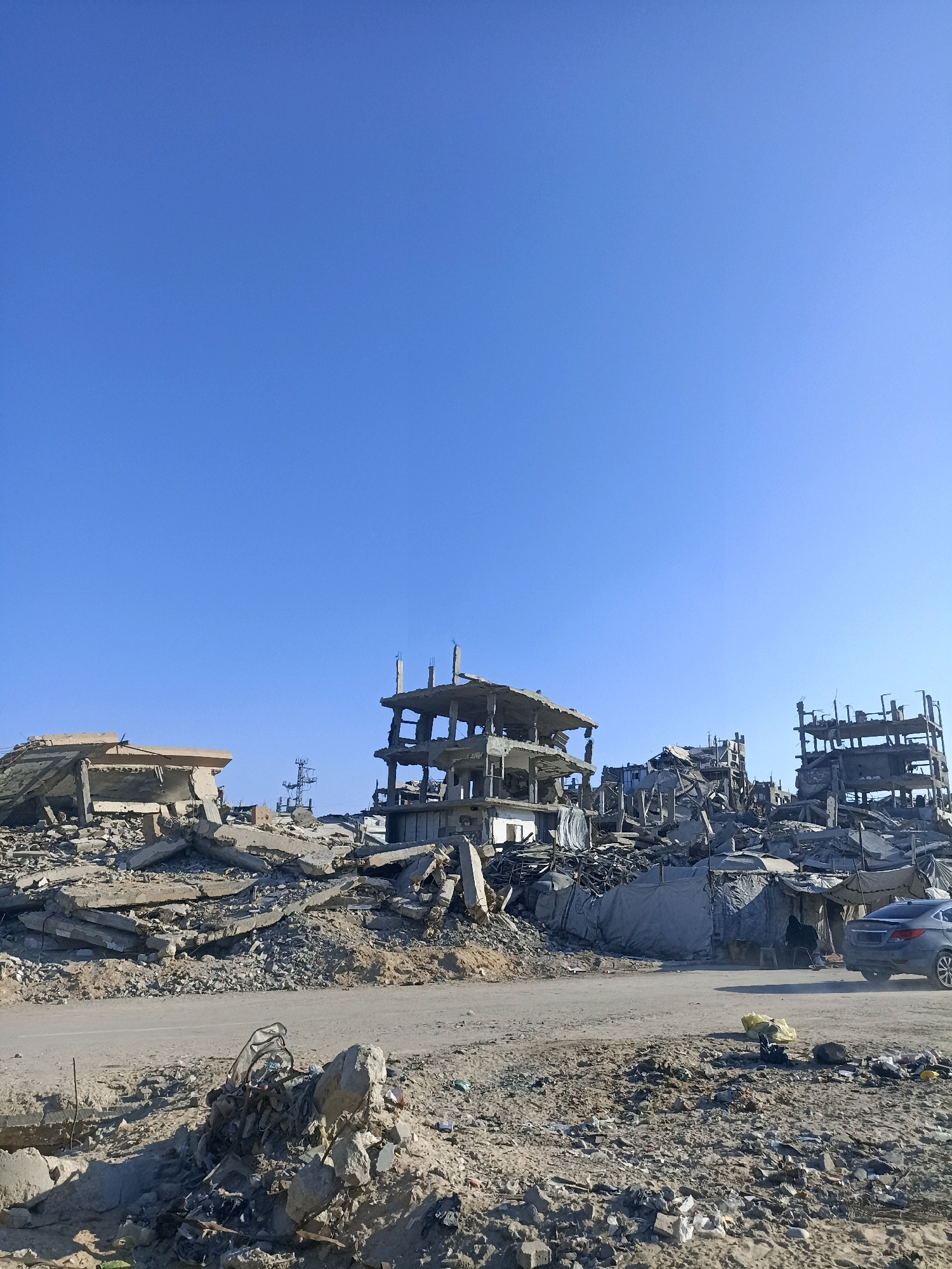 A wide, low-angle shot captures a devastated urban landscape under a clear, bright blue sky. In the foreground, a rough, debris-strewn ground leads to a paved road. Beyond the road, the remains of several multi-story buildings are visible, characterized by skeletal concrete structures, exposed rebar, and piles of rubble. The buildings appear to have been heavily damaged, with only partial walls and floors intact. One structure on the right shows multiple floors with visible cross-beams. A dark-colored sedan is parked on the road to the right, partially obscured by a makeshift shelter constructed from tarps. A single person is visible within the shelter. The overall impression is one of destruction and hardship.