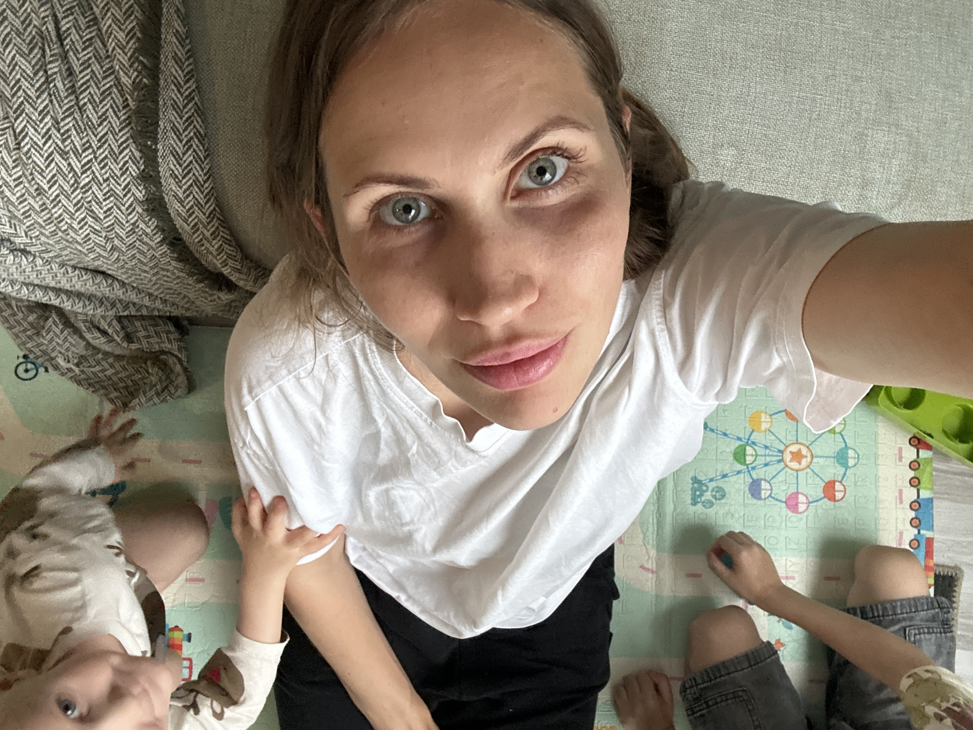 A woman is taking a selfie from a high-angle perspective. She is wearing a white t-shirt and black pants. Two children are visible in the frame, one to the left wearing a white shirt with brown reindeer, and the other to the right wearing grey shorts. They are sitting on a patterned playmat. A patterned grey blanket is visible in the top left corner. The location appears to be inside a home. It is not possible to confirm that the location is in Fethiye, Turkey from the visual data.