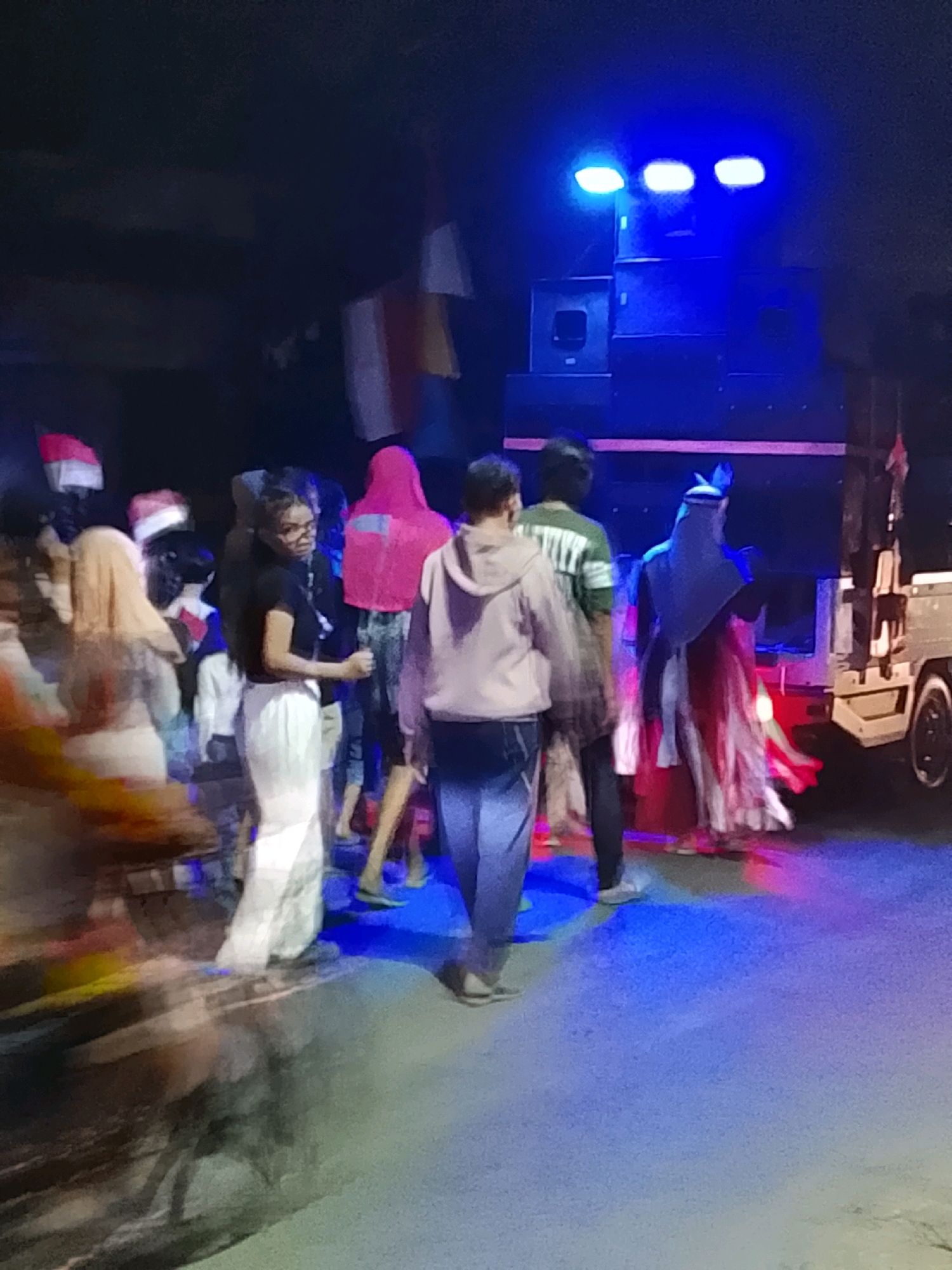 The image depicts an outdoor gathering at night in Ujungrusi, Indonesia, characterized by vibrant blue and pink lighting. A group of people are visible, some appearing to be dancing or socializing. A prominent element is a stack of large speakers, illuminated by blue stage lights from above. Several flags, appearing to be Indonesian flags, are present, some on poles and others held by individuals. The ground is wet or reflective, illuminated by the colored lights. In the background, a vehicle, possibly a truck or van, is partially visible to the right. The overall atmosphere suggests a festive or celebratory event.