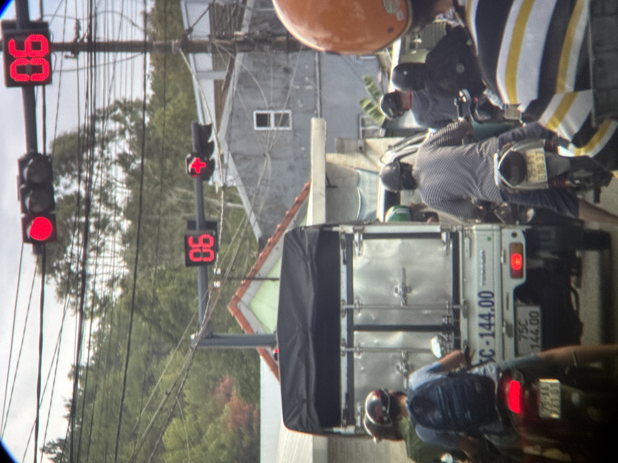 The image depicts a street scene in An Cựu Ward, Vietnam. In the foreground, several individuals on motorcycles are visible. One person in the center is wearing a blue backpack and is on a red scooter. To their left, another person in a green shirt and a light-colored helmet is partially visible. To the right, two individuals are seen on motorcycles, one wearing a dark helmet and the other wearing a brightly colored orange helmet. A white truck with a black tarp covering its bed is stopped behind the motorcycles. The truck has license plate markings that include "SC-144.00" and "75C 144.00". In the background, buildings with distinct architectural features, including tiled roofs, are present. Traffic lights with red lights illuminated are visible overhead, along with a digital countdown timer displaying "06". Power lines crisscross the sky. The overall environment suggests a typical urban street during daytime.