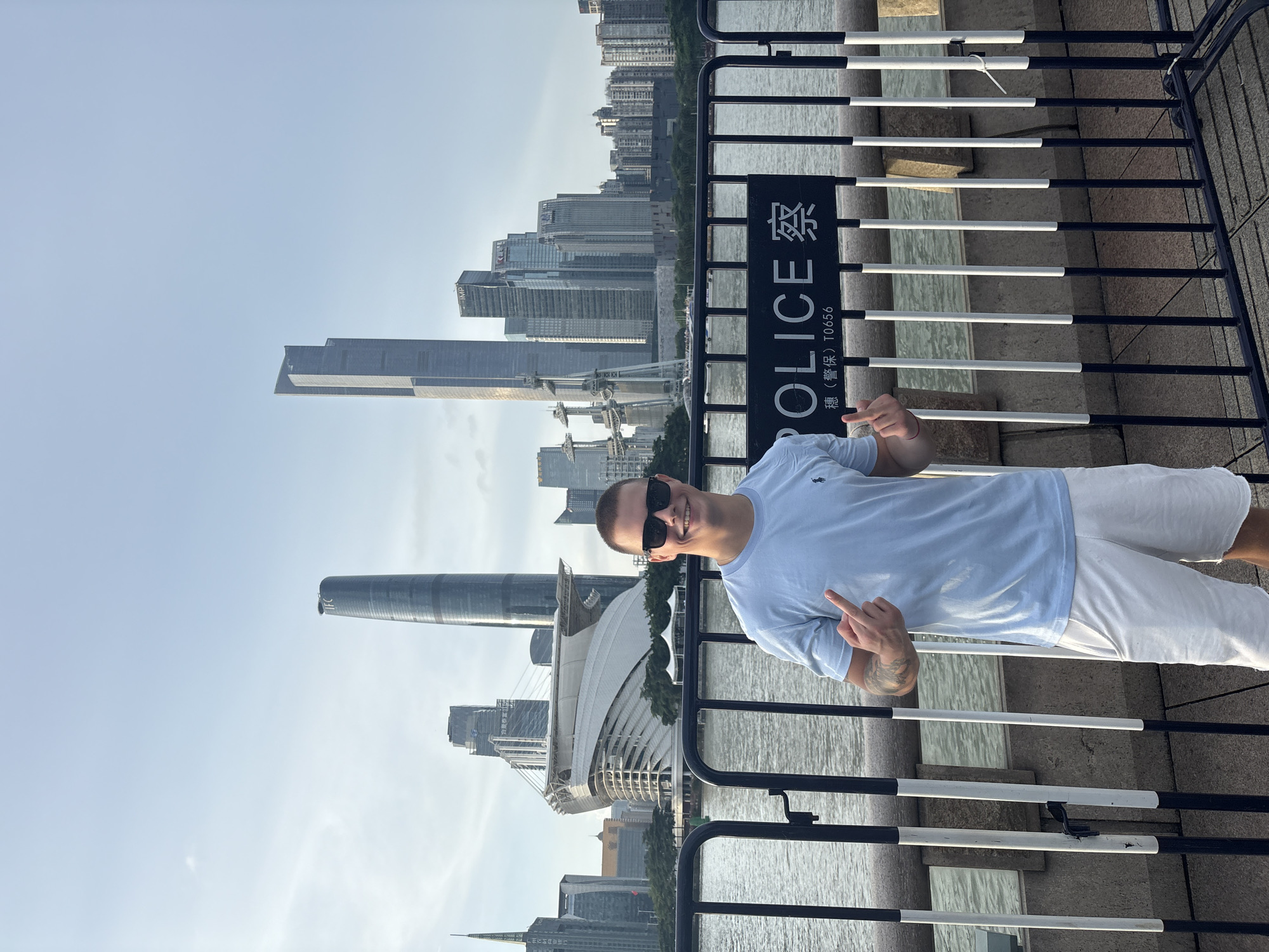 A man stands in front of a body of water with a city skyline in the background, in Haizhu District, China. The man, who is facing the camera and smiling, is wearing sunglasses, a light blue t-shirt, and white shorts. He is making a hand gesture with both hands, with his thumbs up and index fingers pointing up. Behind him is a black metal fence with white and black vertical bars. A sign on the fence reads "POLICE" in English and Chinese characters. The cityscape features several tall modern buildings, including the Canton Tower, and a large, white, futuristic-looking stadium or arena. The water appears calm, and the sky is a light blue with some clouds.