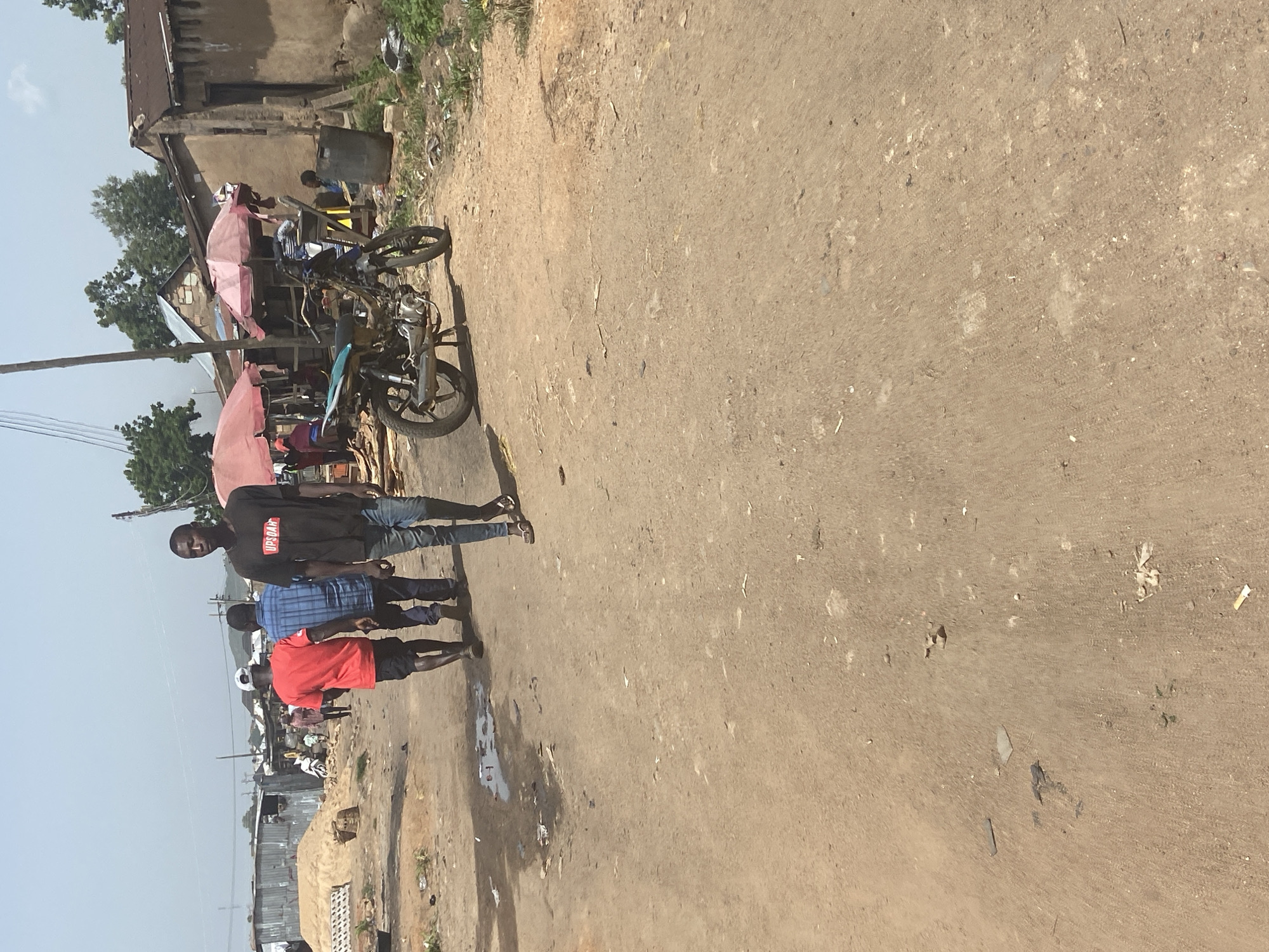 The image depicts a bustling street scene in Bukuru, Nigeria, likely a market area. In the foreground, a dirt road is visible, marked with puddles and scattered debris. Several individuals are walking along the road. A young man in a black t-shirt with "UPSOAR" printed on it is walking towards the viewer, followed by two other men, one in a red t-shirt and the other in a blue checkered shirt. A motorcycle is parked on the side of the road, in front of market stalls with pink umbrellas. The background reveals a collection of buildings constructed with corrugated iron and mud, characteristic of informal settlements or market structures. Trees and a bright sky complete the scene, suggesting a warm, sunny day. The overall impression is one of everyday life and commerce in a developing urban environment.
