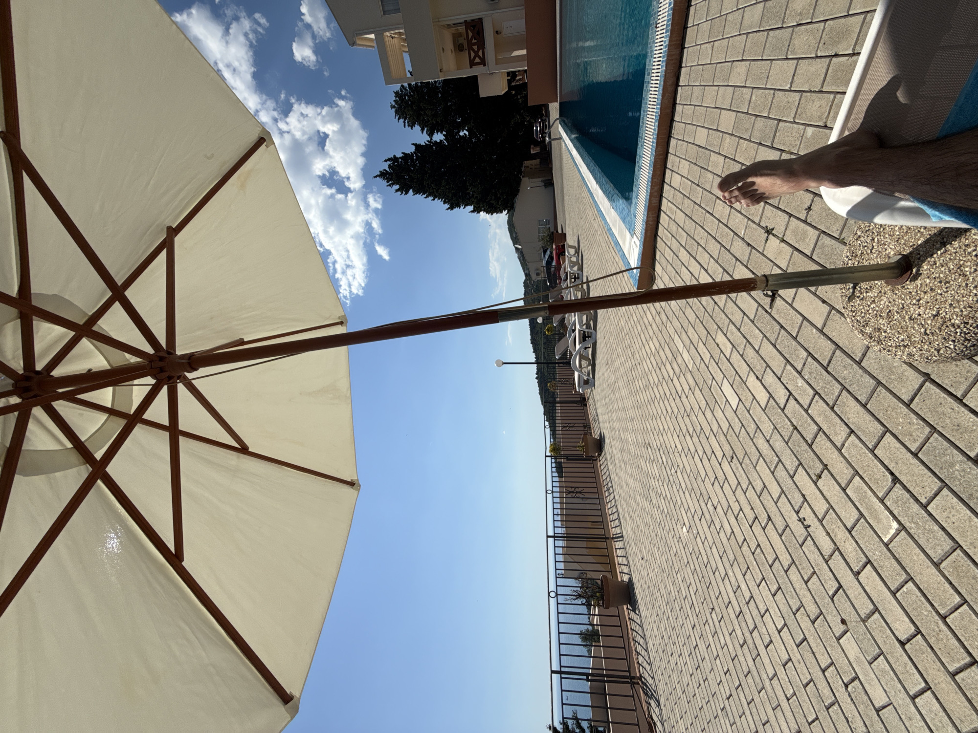 The image depicts a scene in Šušanj, Montenegro, near a swimming pool. The camera captures the underside of a white parasol in the foreground, with wooden support beams visible. A person's leg and foot are seen reclining on a sunbed next to the pool's edge. The pool is tiled with blue and white tiles, and is placed directly besides a brick walkway. The walkway is paved with light-colored bricks and extends into the distance. Beyond the pool, a dark railing surrounds the outdoor area. Farther away, there is a glimpse of the sea, a distant landscape with buildings, street lamps, and trees. The sky is blue with scattered clouds. The picture is taken during the day.