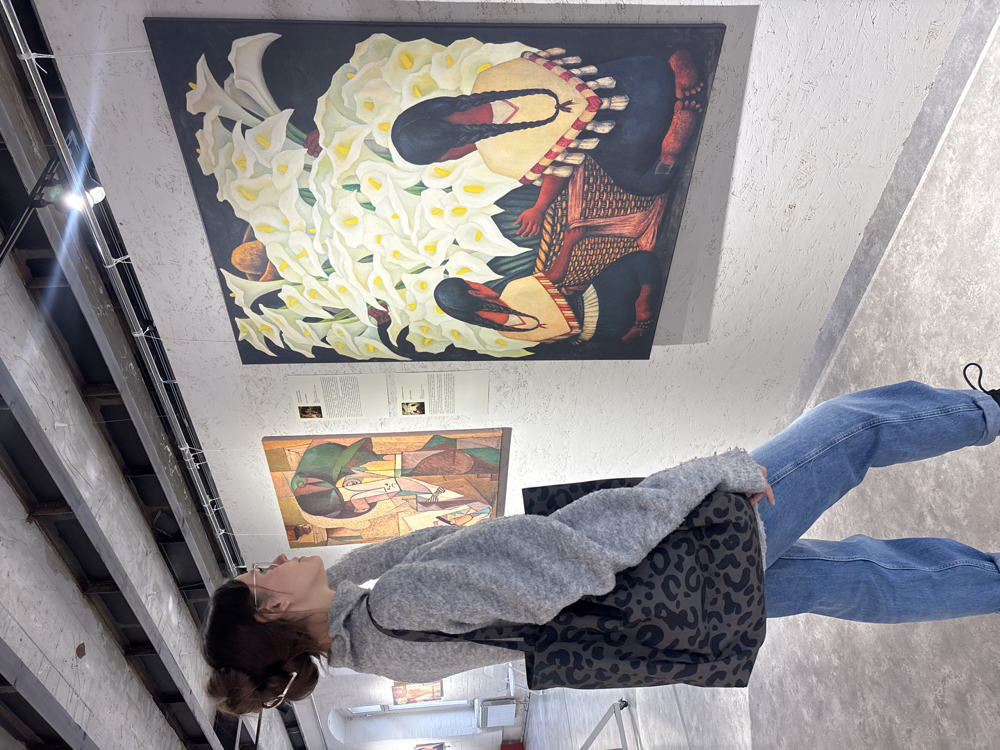 A young woman stands in what appears to be an art gallery or museum in Krasnoyarsk, Russia. She wears jeans, a gray sweater, and a black animal print purse. She looks upward at a large painting of white lilies with two women dressed in indigenous garb, positioned above another painting of stylized figures. The environment is a modern space with an exposed ceiling and support beams.
