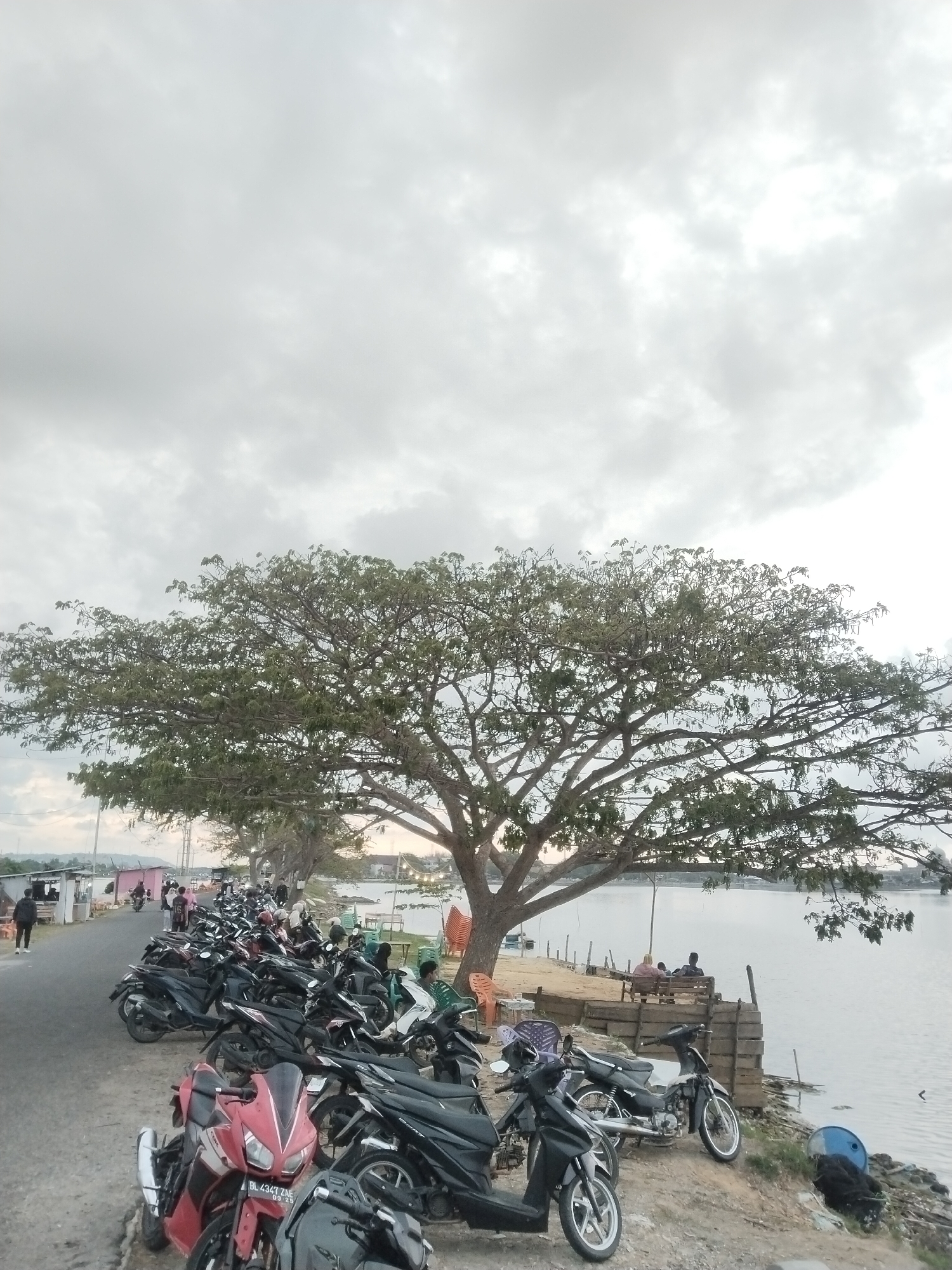 The image shows a waterfront scene in Lhokseumawe, Indonesia, under an overcast sky. A large tree dominates the foreground, its branches extending widely. A row of parked motorcycles lines a paved road adjacent to the water's edge. Several people are visible: some are walking along the road, while others are seated on benches near the water. A partially constructed wooden platform extends toward the water, with people sitting on a bench located there. Various objects are scattered near the water, including chairs, plastic containers, and discarded items. The water appears calm.