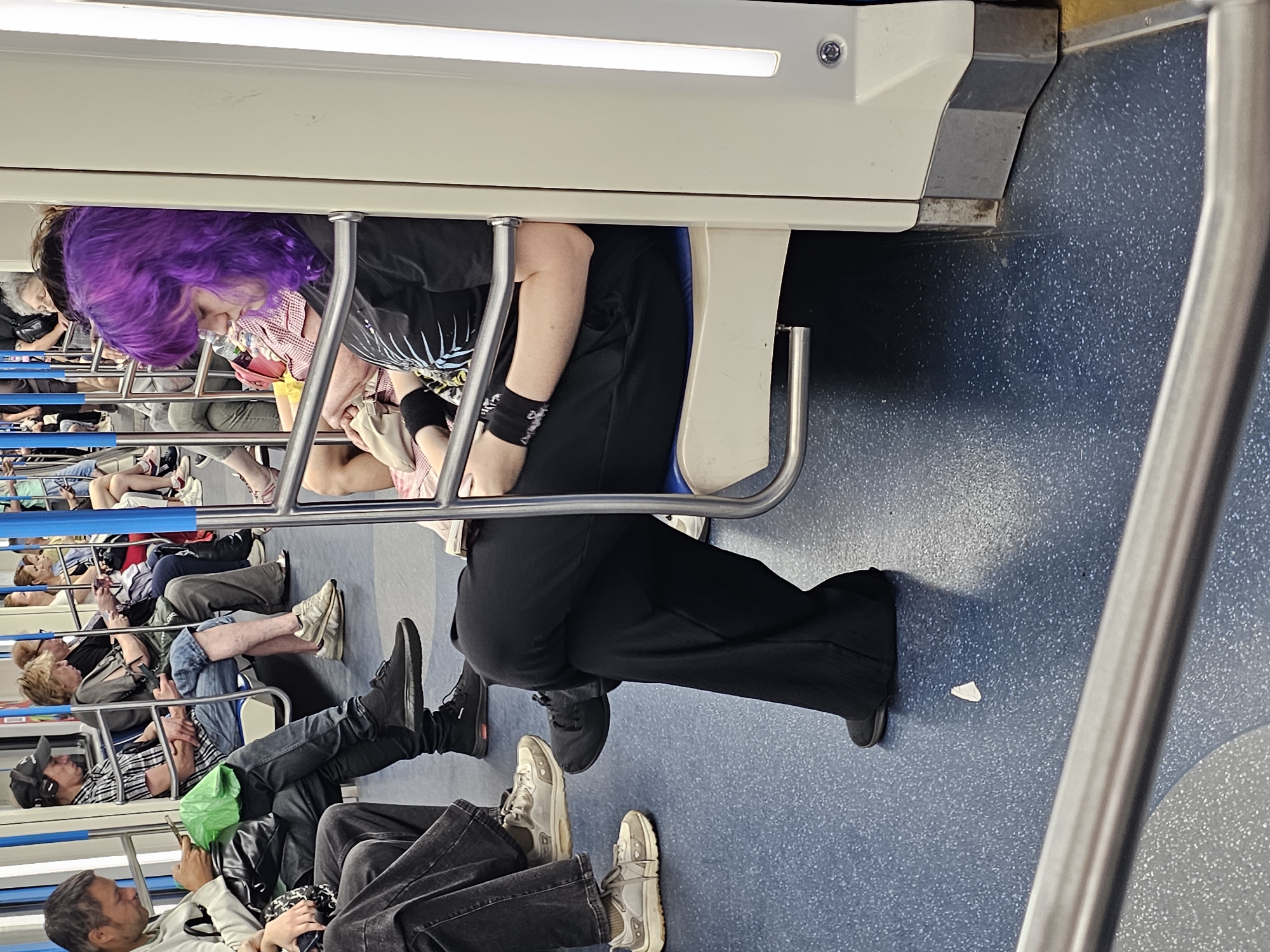 A medium shot captures several individuals inside a subway car. The floor is a speckled blue, and vertical poles are visible throughout the scene. In the foreground, a person with vibrant purple hair is seated, wearing a black t-shirt with a graphic print and black pants. They are looking down, possibly at a phone. To their left, another person is seated, wearing dark denim jeans and a light-colored hoodie. They are also looking down at a phone. Further to the left, a man in a black baseball cap and plaid shirt is seated, wearing headphones and looking at his phone. Behind him, other passengers are visible, some looking at their phones, others seated or standing. The overall impression is of a typical commute, with passengers occupied with their devices.