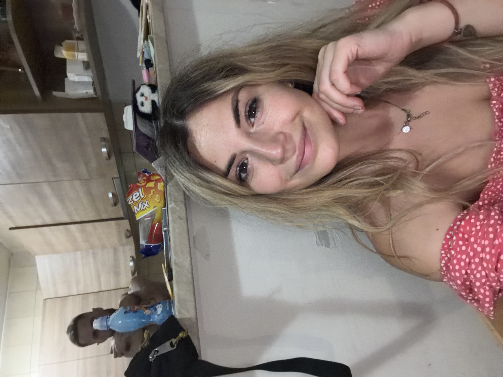 A young woman with blonde hair and fair skin is captured in a close-up selfie, smiling and resting her chin on her hand. She is wearing a pink polka-dot off-the-shoulder top and a necklace with a circular pendant. Her hair is long and wavy, falling around her shoulders. The background appears to be a kitchen, with wooden cabinets and shelves visible. A man, partially blurred, can be seen in the background to the left, near a blue bottle. Several items are scattered on the counter, including a bag of snacks and a small panda figurine. The overall impression is a casual, indoor setting, possibly a home kitchen in Budva, Montenegro.