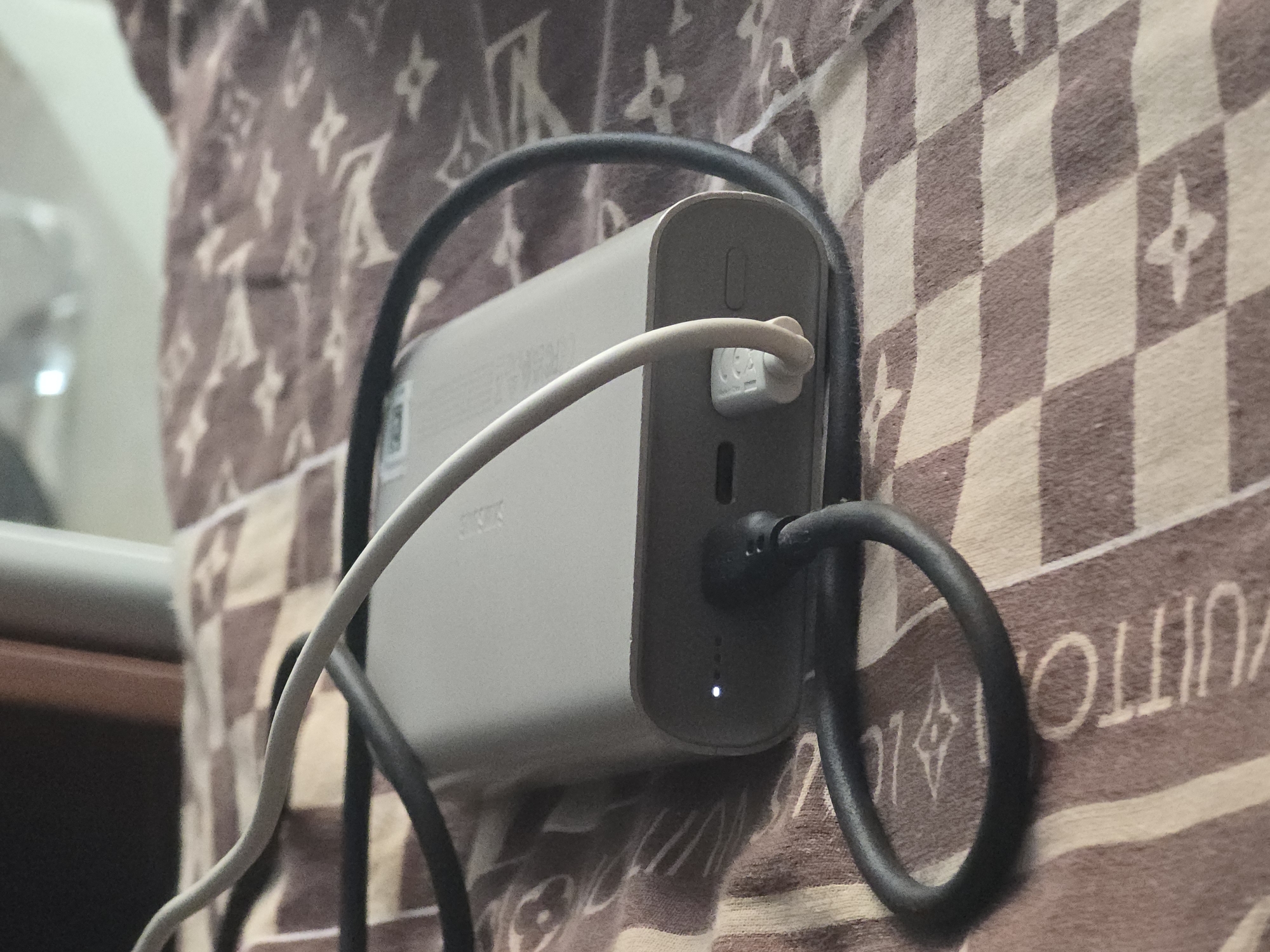 The image shows a close-up of a light gray power bank on a patterned blanket. Two charging cables are connected to the power bank. One cable, which appears to be black, is looped around the power bank. The other cable, which is white, is plugged into the power bank and leads out of the frame. A small white LED light is illuminated on the power bank, indicating it is charging or powered on. The blanket has a repeating checkered pattern with a floral or star-like motif. The background is blurred, but appears to show a dark object and a lighter, out-of-focus area.
