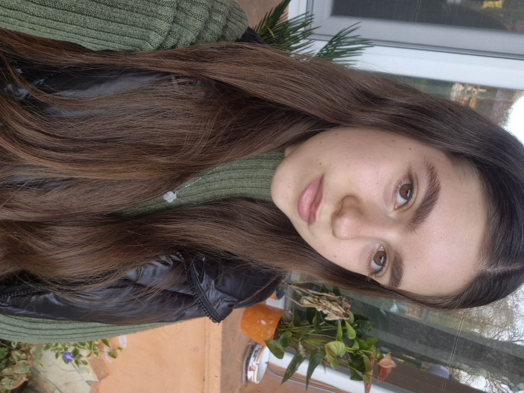 A young woman with long, brown hair is posing for a selfie-style photo. She has fair skin, brown eyes, and well-defined eyebrows. She is wearing a green sweater and a black, puffy jacket. A silver necklace is visible on her chest. The background shows an exterior setting with greenery, including a plant in an orange pot. The photo was taken near a window, reflecting outdoor scenery. While it is impossible to verify the precise location is in Pavlikeni, Bulgaria, there are architectural elements and foliage that are in line with that part of the world.