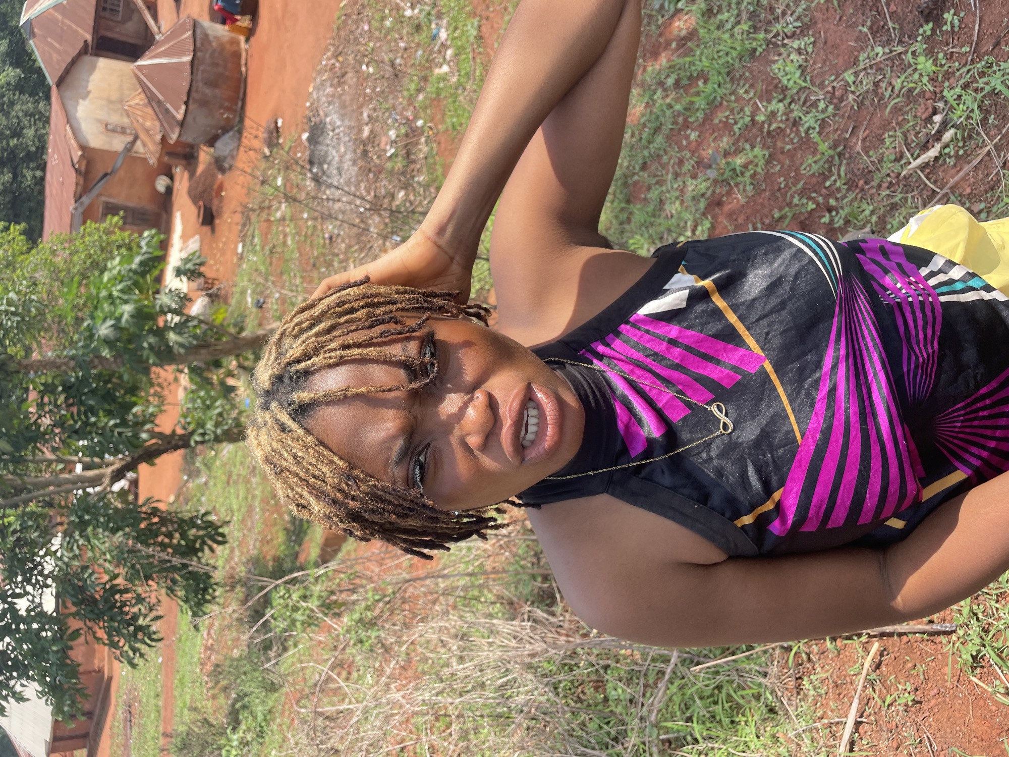 A young Black woman is prominently featured in this vertically oriented photograph, captured outdoors in what appears to be a rural or semi-urban setting in Ogbe, Nigeria. She is likely reclining or lying down, looking directly into the camera with an engaging, somewhat playful expression, her mouth slightly open to reveal her teeth. Her right arm is raised, with her hand near her head, possibly adjusting her hair or posing.
She is wearing a black sleeveless top with a vibrant geometric pattern of fuchsia pink, black, and gold lines and stripes. A delicate golden necklace with an infinity symbol pendant is visible around her neck. Her hair is styled in bleached or light-colored dreadlocks. Part of a yellow garment or item can be seen to her lower right.
The background shows a mix of natural elements and simple architecture under bright daylight. Buildings with rusted, corrugated metal roofs and light-colored walls are discernible behind her, characteristic of local housing. The ground is covered with reddish-brown soil, sparse green grass, and dry vegetation, suggesting a warm and possibly arid or dry season environment. Green foliage and trees are also visible, adding depth to the scene. The image conveys a candid moment within a typical Nigerian landscape.
