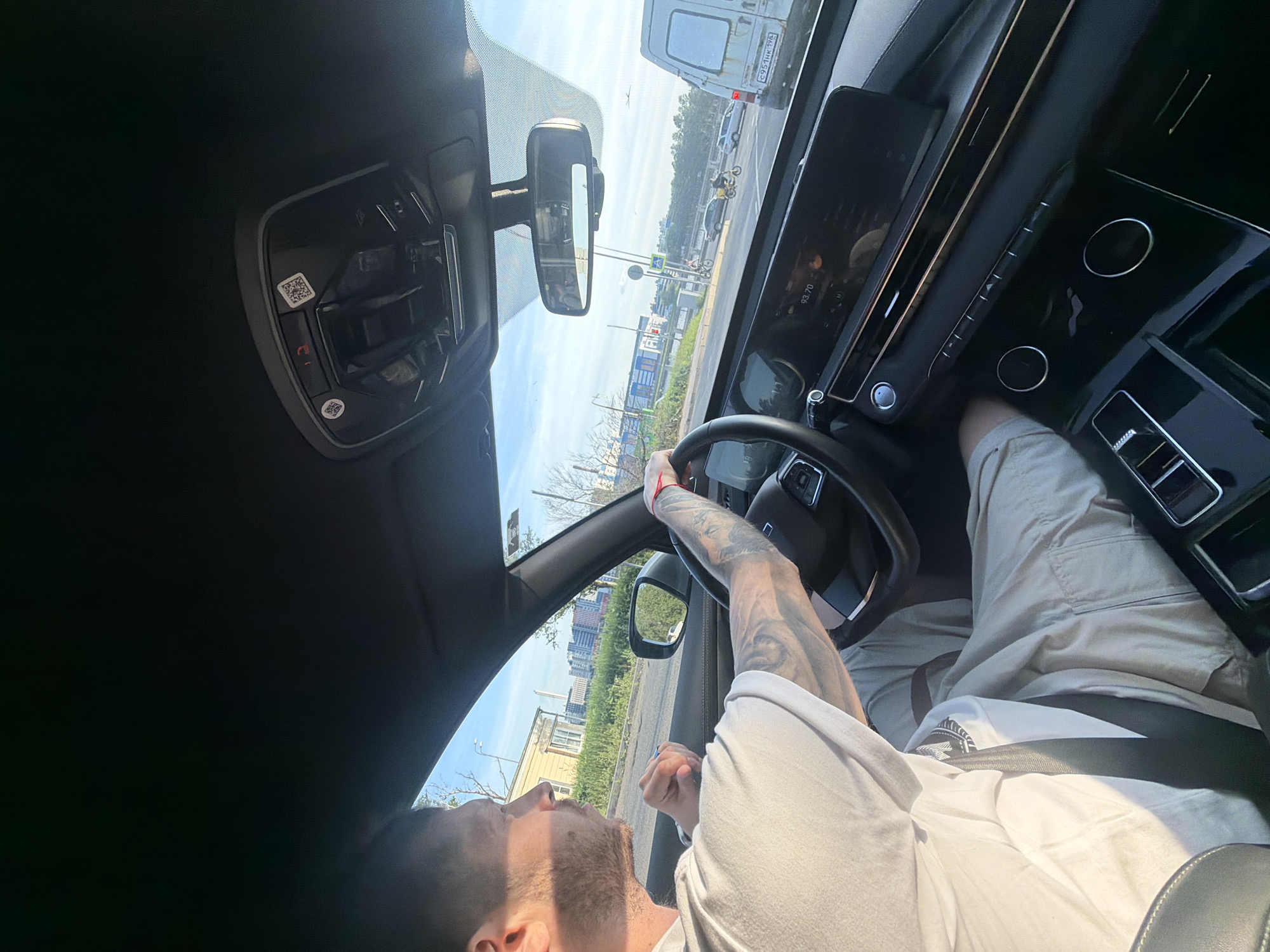 The image captures the interior of a vehicle from the driver's side, situated in Saint Petersburg, Russia. A male driver, with a beard and tattoos on his left arm, is visible, wearing a light-colored t-shirt and shorts, secured by a seatbelt. His hands are positioned on the steering wheel. The vehicle's interior features a modern digital dashboard displaying "93.70," a large central infotainment screen, and an overhead console with an SOS button and two QR codes. Through the windshield, an urban street scene is visible under a clear blue sky. Traffic includes a white panel van with a Russian license plate "C951HK 198" (Saint Petersburg region code), a dark sedan, a yellow vehicle, and two individuals on e-bikes on the road. A pedestrian crossing and a traffic light showing red are also present. The background reveals an older wooden building, modern multi-story buildings, and distant industrial structures.