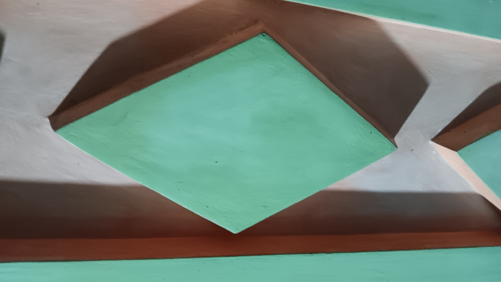 The image shows a close-up view of a textured wall with geometric patterns. The main focus is a large, light green diamond shape that is raised from the wall, casting a shadow. To the left of the diamond, a narrow, vertical light green rectangle is visible. To the right and below the diamond, another light beige geometric shape is partially visible, also casting a shadow. The background wall appears to be a light, neutral color with subtle variations in tone due to lighting. The lighting appears to be coming from the upper right, creating distinct shadows that emphasize the three-dimensional nature of the geometric elements. The overall impression is of a decorative architectural feature or a section of a building's facade. The location is stated to be in the city of Sonobekel, Indonesia.