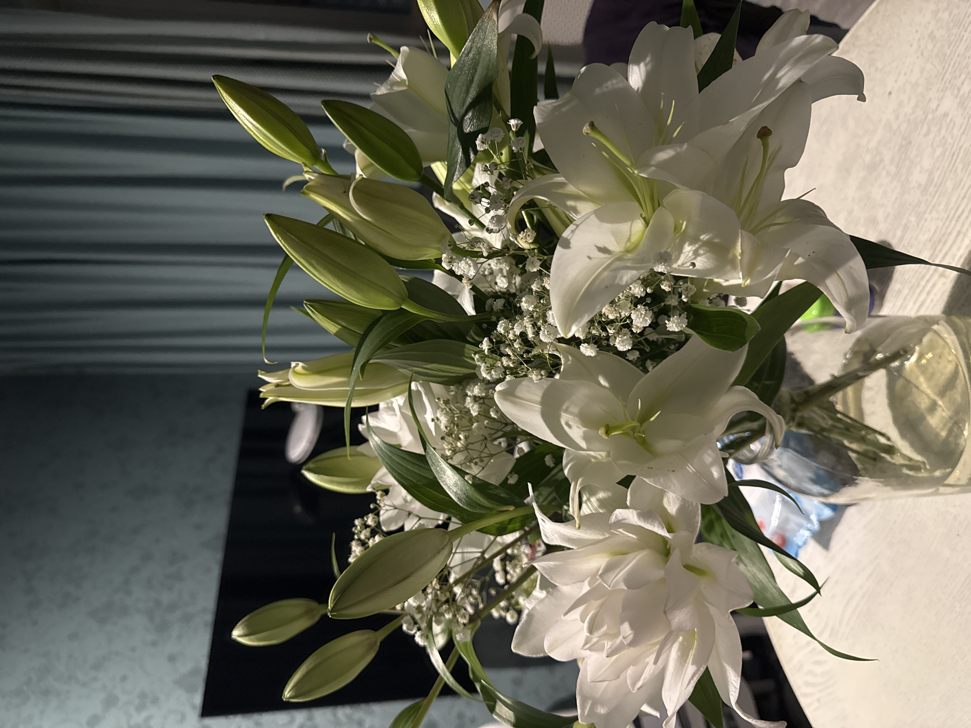 A bouquet of white lilies and baby's breath is arranged in a clear glass vase. The bouquet features a mix of open blooms and closed buds, with green leaves providing contrast. The flowers are situated on a light-colored surface. In the background, a dark, striped surface is visible. The location is Unknown, Unknown.
