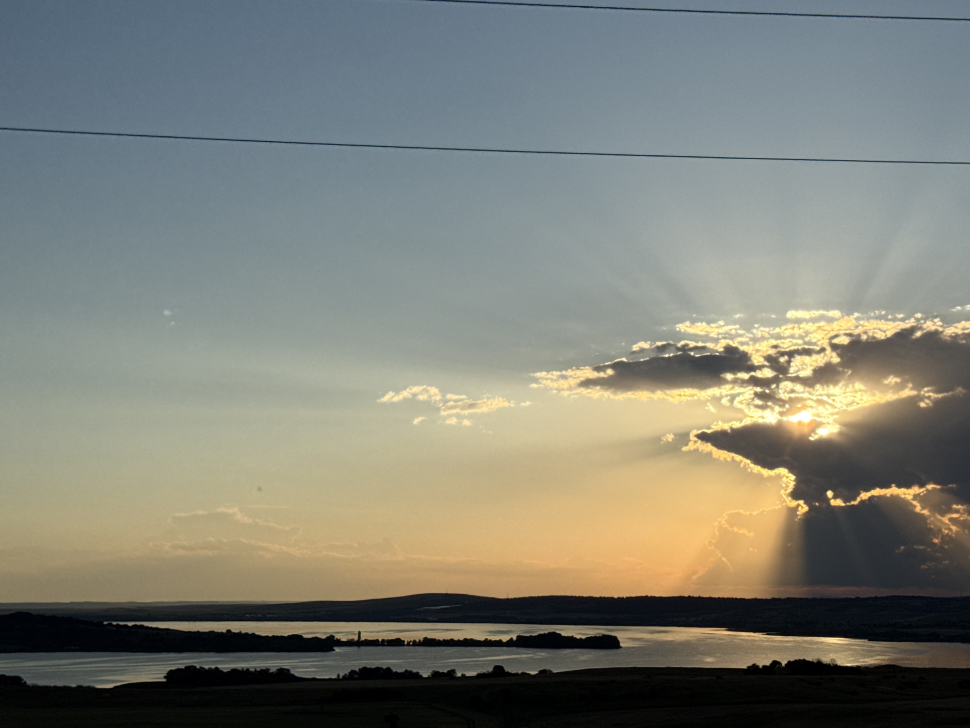 The image captures a serene natural landscape at sunset, likely near the city of Tvarditsa, Bulgaria. A vast body of water, possibly a lake or reservoir, dominates the lower half of the frame, reflecting the warm hues of the setting sun. Silhouetted against the water are dark, rolling hills covered in vegetation, their shapes softened by the low light. In the distance, a thin strip of land is visible, dotted with dark trees and a single, tall, slender structure that could be a tower or a lighthouse. The sky above transitions from a soft, pale blue near the top to a vibrant orange and yellow glow near the horizon, where the sun is partially obscured by dramatic, dark clouds. Rays of light pierce through these clouds, creating a breathtaking sunburst effect. Two thin, dark power lines stretch horizontally across the upper portion of the sky, suggesting the presence of infrastructure near this otherwise untouched natural setting. The foreground is a dark silhouette of rolling hills, adding depth and framing the scene.