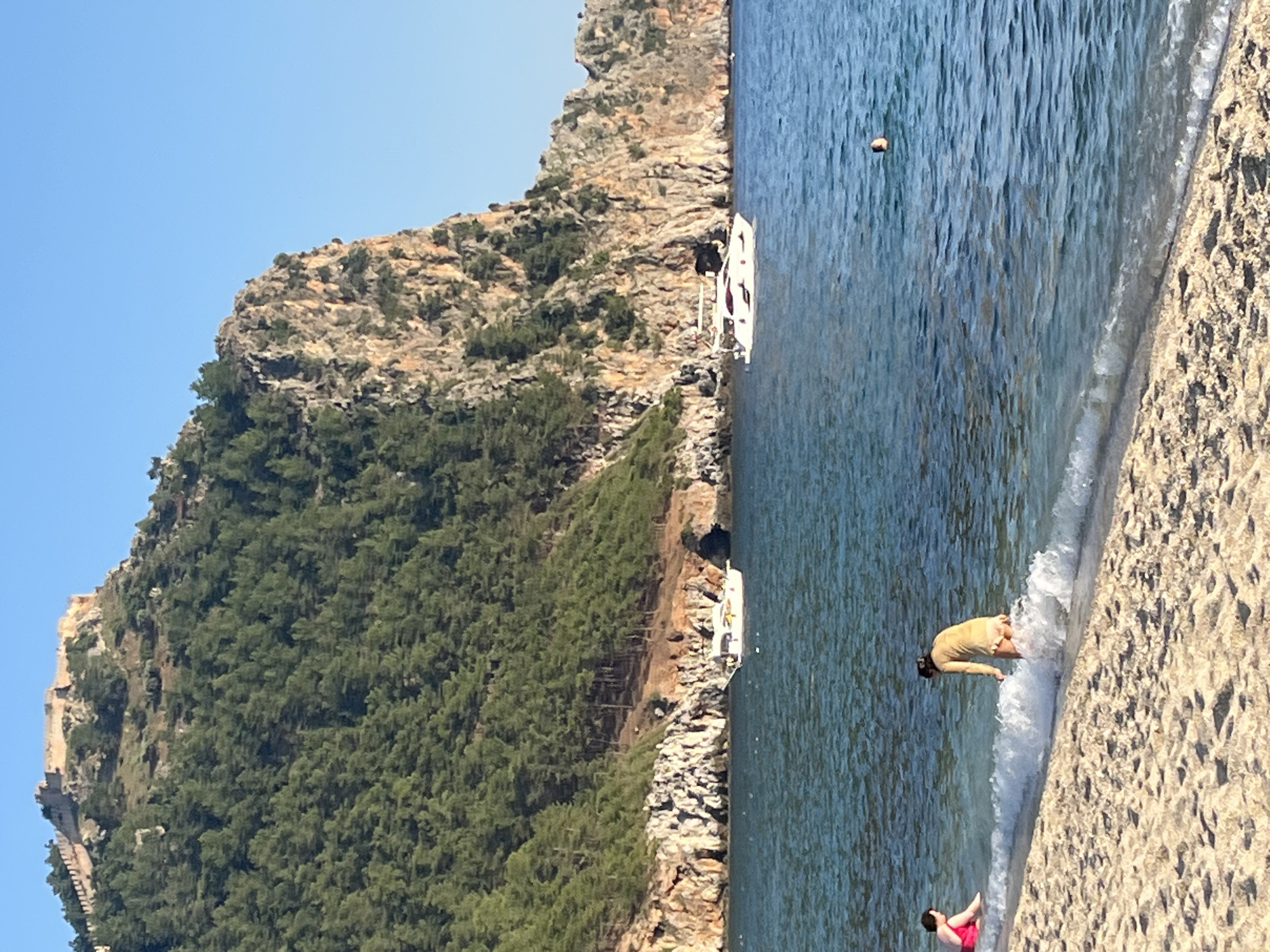 The image depicts a coastal scene in Alanya, Turkey. The view includes a beach, the sea, and a large, rocky, tree-covered hill topped with the Alanya Castle fortress. Two boats are visible near the base of the hill along the coastline. Two people are in the water near the beach. One person is bent over, immersed in the shallows, while another individual is in a red swimsuit. The water appears calm and clear. The sky is clear and blue.