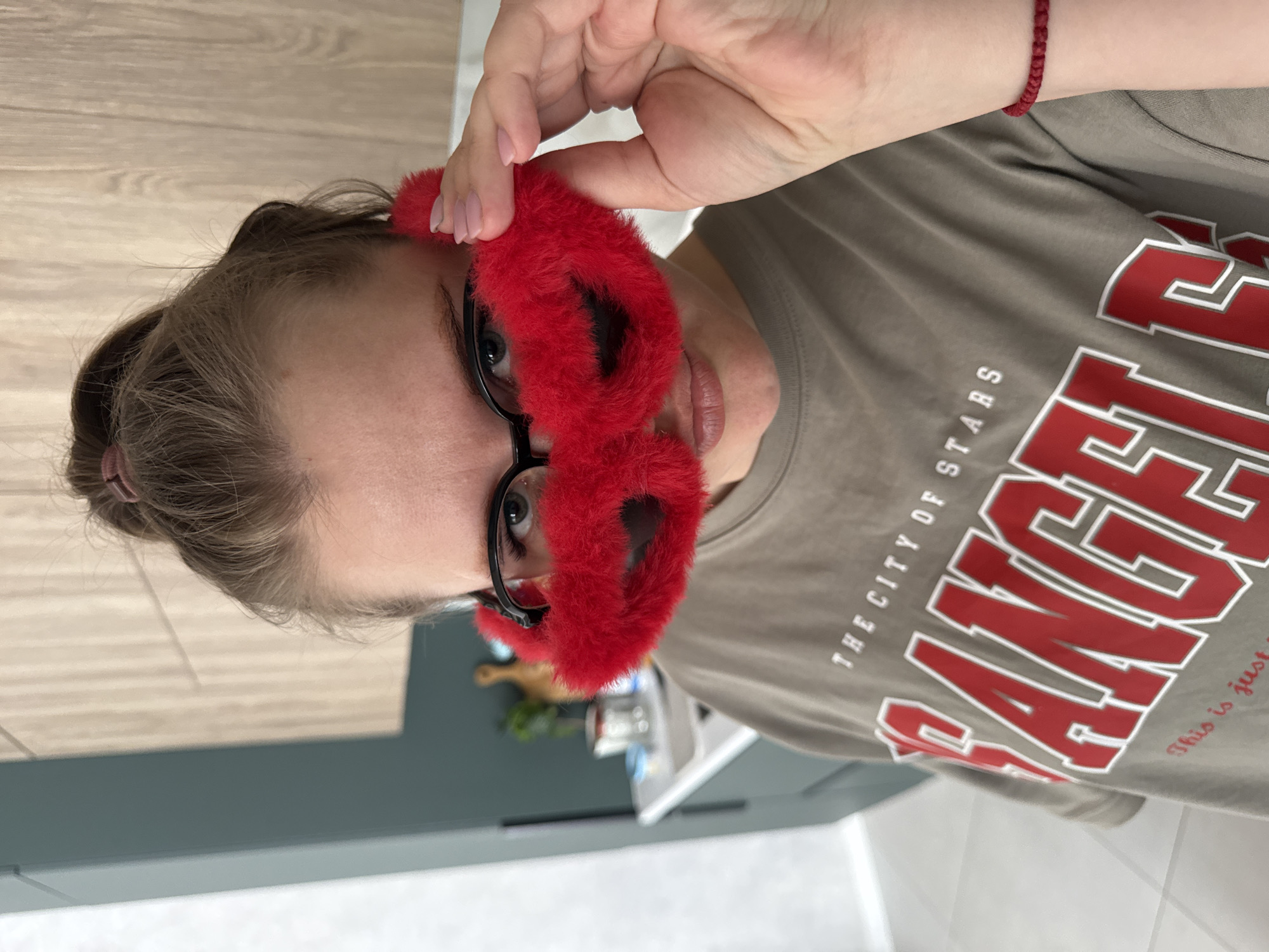 A woman is holding a red, fuzzy mask up to her face, obscuring the area around her eyes. She is wearing glasses with black frames. The woman has light brown hair pulled back in a bun. She is wearing a grey t-shirt with large, red lettering that reads "LOS ANGELES THE CITY OF STARS". A red bracelet is visible on her left wrist. The background includes a light-colored countertop, a dark grey or green wall, and some kitchen items. The location is Gornyi (Dvurechenskoe S/P), Russia.