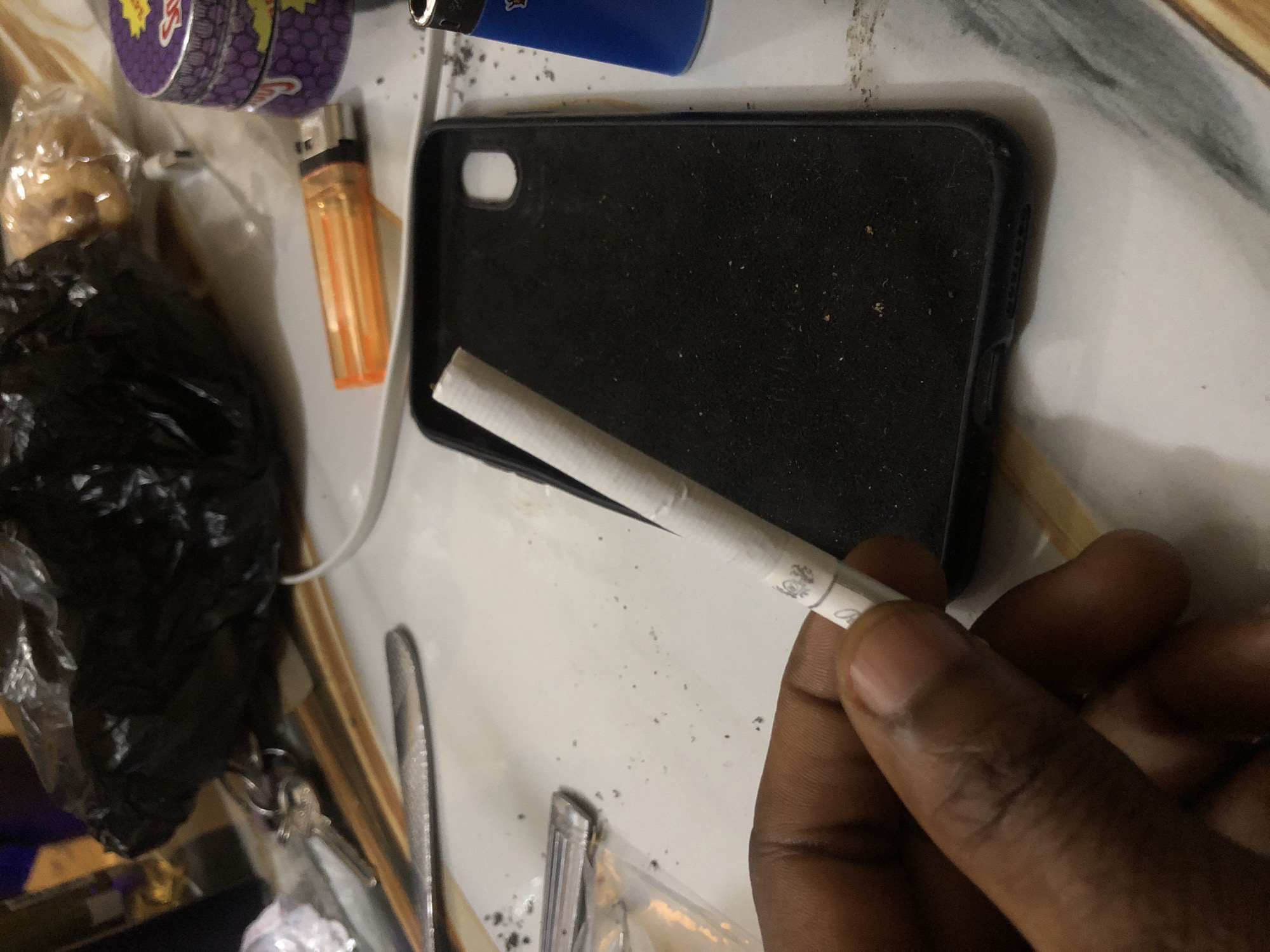 The image depicts a close-up view of a hand holding a partially rolled cigarette. The background reveals a cluttered surface with various objects, including a black phone case, an orange lighter, and a purple metal container. Scattered particles, possibly tobacco or ash, are visible on the surface. The lighting suggests an indoor environment. There are no indications of the location being in Kabong, Nigeria.