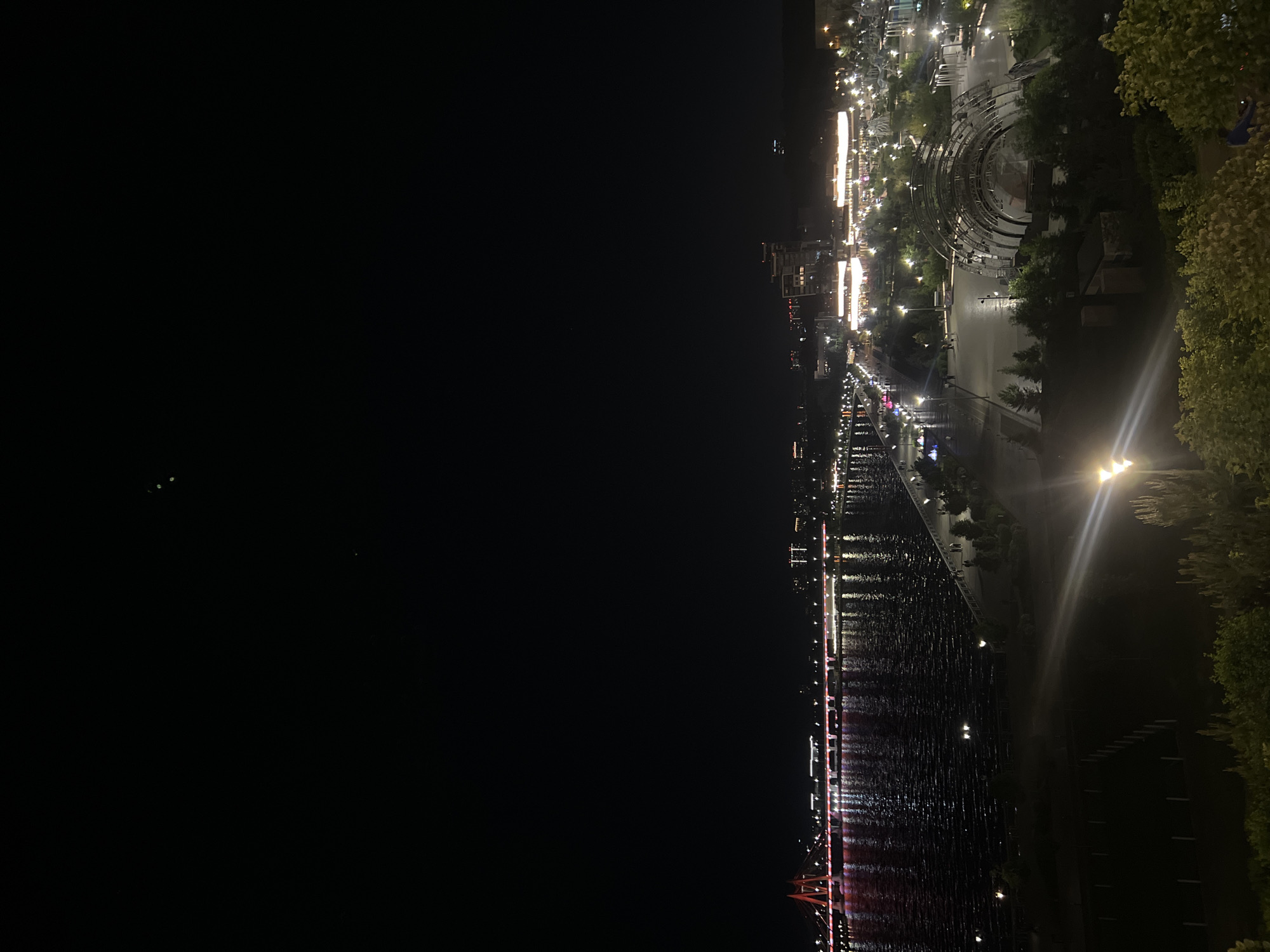 The image depicts a night scene in Kazan, Russia, capturing the cityscape along the Kazanka River. A dark expanse of water dominates the lower half of the frame, reflecting the city lights. The river is spanned by a bridge, visible at the bottom of the image, its structure illuminated with red lights. The cityscape extends along the riverbank, with streetlights, buildings, and infrastructure clearly visible. Prominently displayed are a Ferris wheel, and the Kazan Family Center. The photo was taken from an elevated vantage point from the opposite side of the river, with trees and greenery at the edge of the composition.