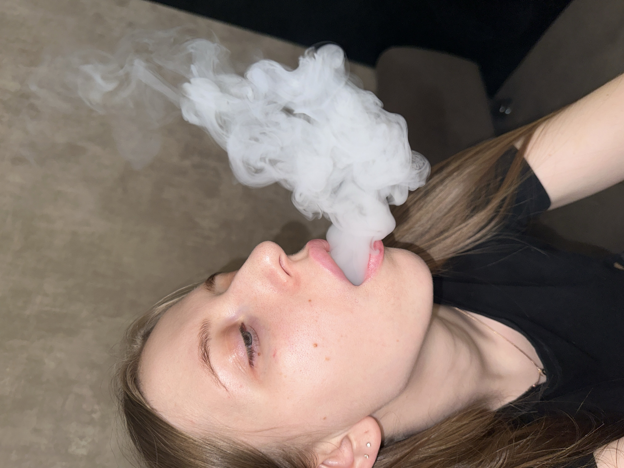An indoor close-up photograph, taken in Chaikovsky, Russia, features a young woman in profile exhaling a dense white plume of vapor. The woman, with fair skin and light brown hair, is positioned with the right side of her face prominently displayed. Her lips are slightly open and rounded as the vapor exits her mouth. She wears two small, silver-colored stud earrings in her visible earlobe and a thin, light-colored chain necklace over a dark, likely black, t-shirt. Her eyes are partially visible, directed downwards. The background consists of a textured, light brownish-gray wall on the left, transitioning to a dark, blurred area on the right, suggesting an interior setting.