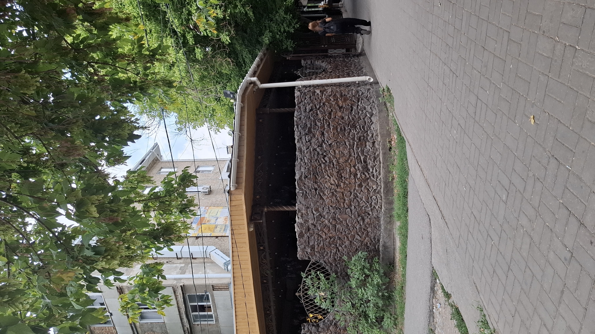 The image shows a street scene in Shymkent, Kazakhstan, with a focus on a building with a stone-clad base and a wooden-roofed veranda. A person with long, reddish-brown hair, wearing black clothing and a backpack, is walking away from the camera on a paved sidewalk. The building appears to be a commercial establishment, possibly a cafe or restaurant, with decorative metalwork visible under the veranda. Adjacent to the building is a multi-story residential building. Lush green trees overhang the scene, their branches laden with seed pods, creating a dappled light effect. The ground is paved with interlocking gray bricks. The overall impression is an outdoor urban environment on a bright day.