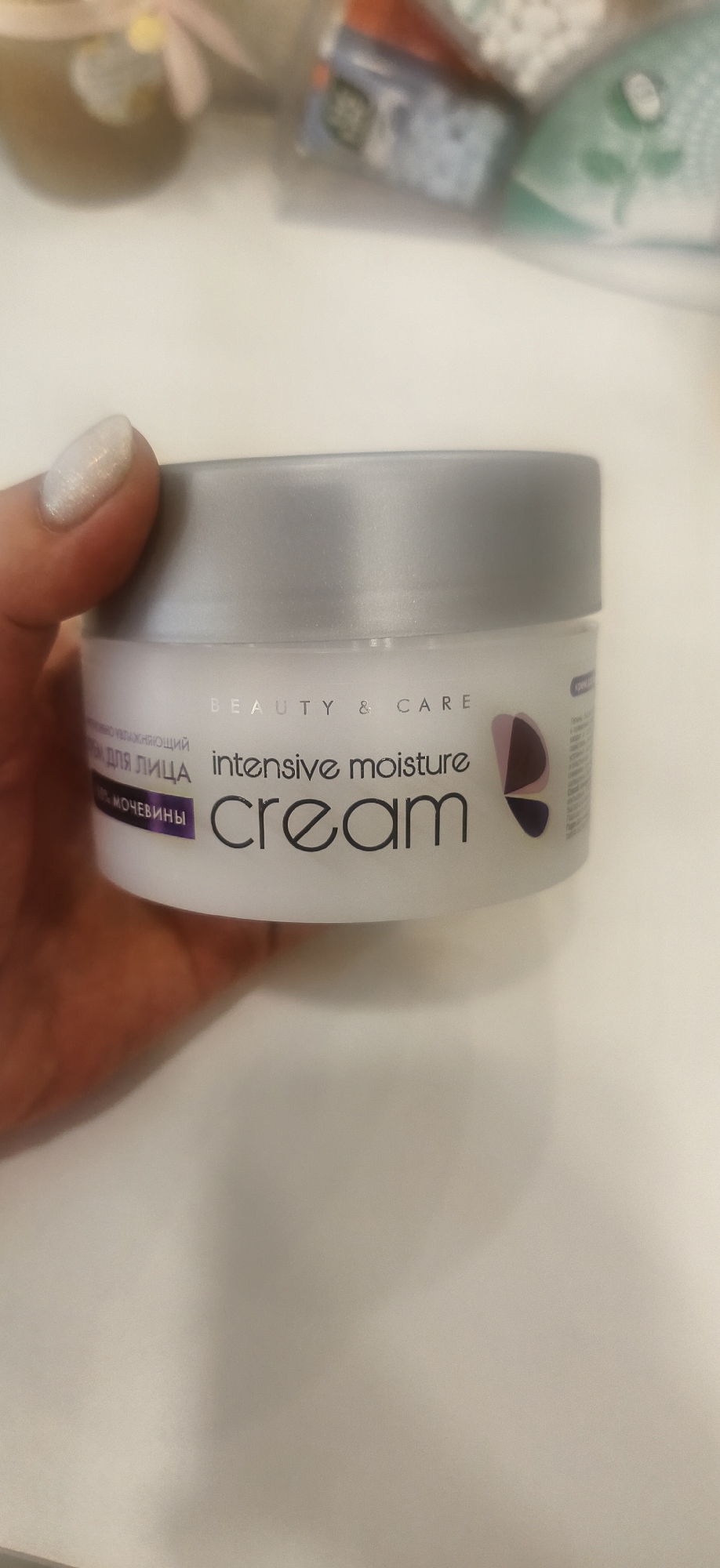 A hand holds a white jar of "Beauty & Care intensive moisture cream." The jar has a silver lid. The background is blurred and shows assorted objects. The location is Unknown, Unknown.