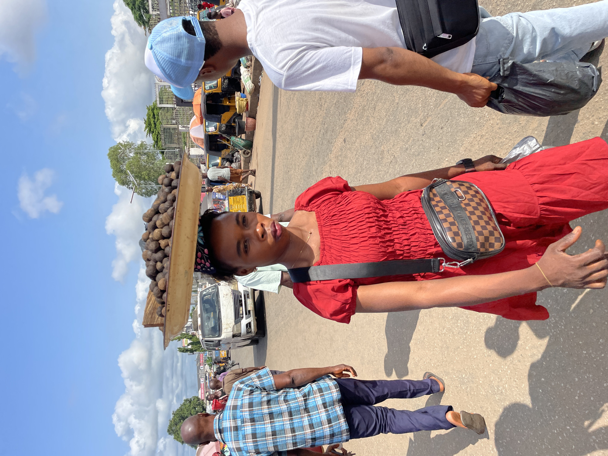 The image depicts a bustling street scene in Onitsha, Nigeria, likely a marketplace given the presence of vendors and vehicles. A young woman, the central figure, is walking forward, balancing a large tray filled with dark, round objects, possibly nuts or fruits, on her head. She wears a vibrant red dress and a black crossbody bag with a checkered pattern. To her right, a young man in a white t-shirt and blue cap walks away from the camera, carrying a black plastic bag. Behind them, other individuals and vehicles, including a white van and yellow auto-rickshaws, are visible. The ground is paved, and the sky is partly cloudy, suggesting it is daytime. The atmosphere appears active and typical of a busy urban environment in Nigeria.