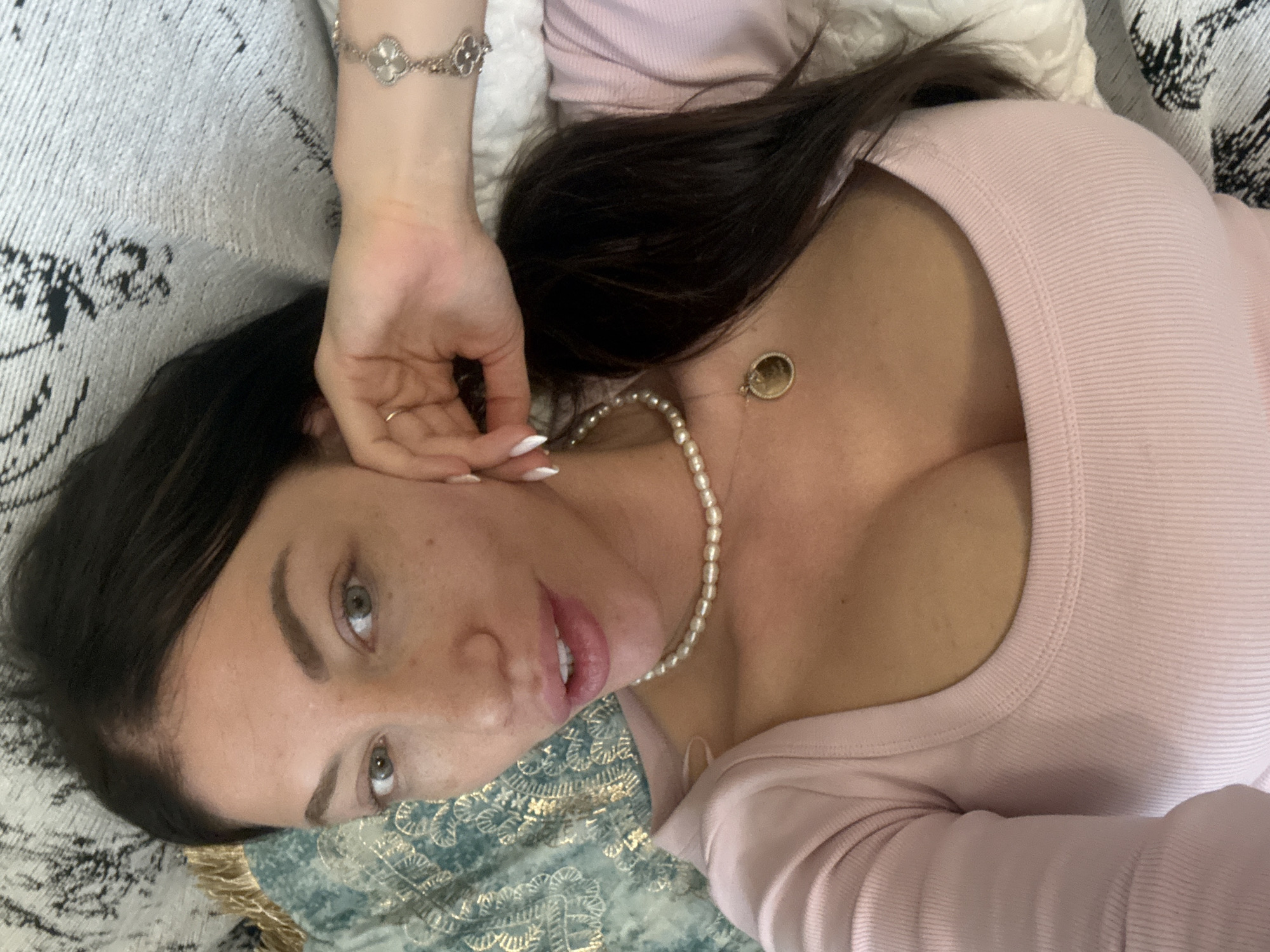 A woman with dark hair and light skin is lying on a patterned surface, wearing a light pink top and a pearl necklace. Her left hand, adorned with a bracelet and a ring, is near her face. A gold pendant hangs near her chest. The location is indeterminate without further information; there's no evidence in the image to specifically place it in Sofia, Bulgaria.