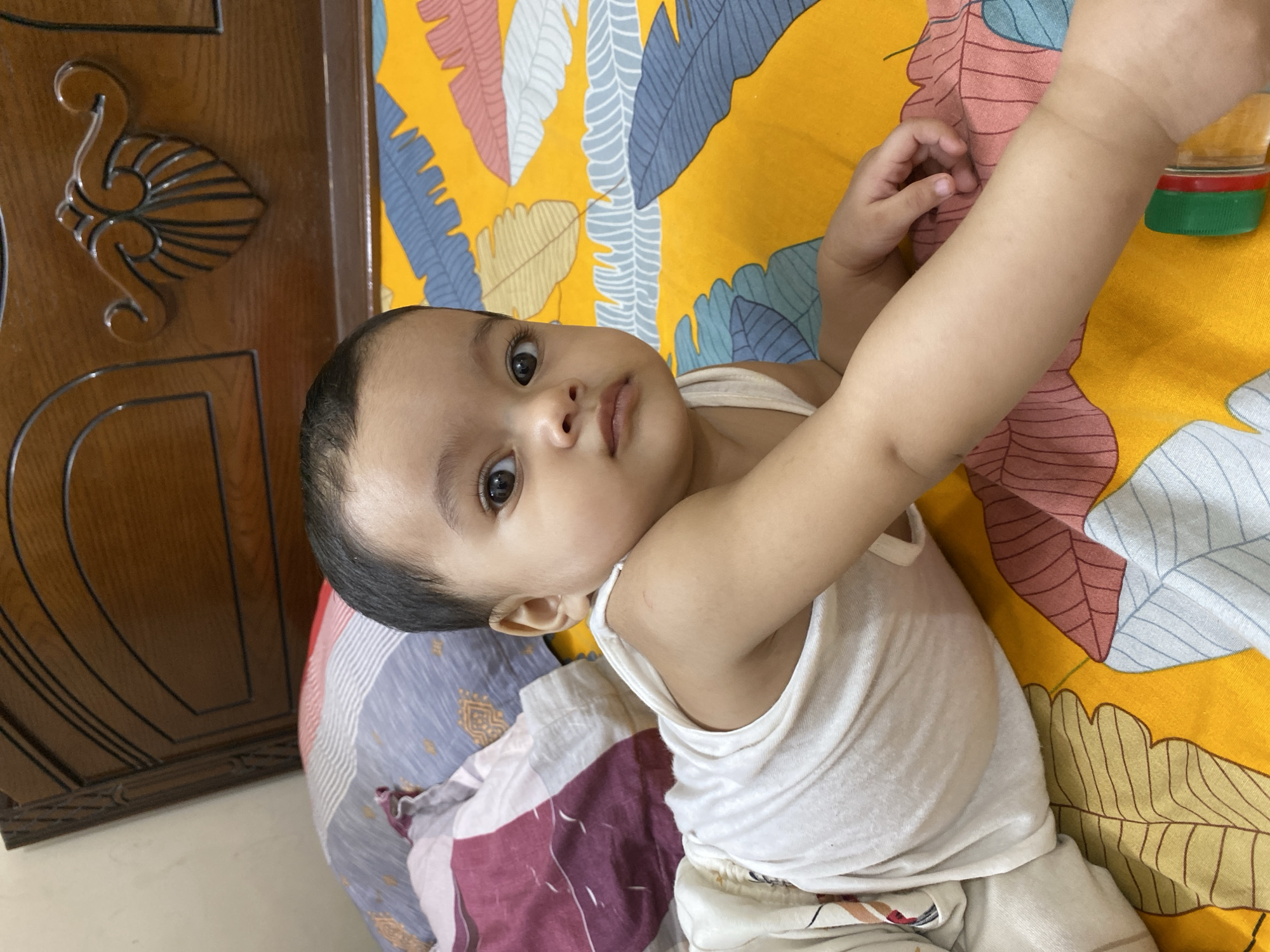 A close-up, high-angle shot captures a baby lying on a bed indoors. The baby, with dark eyes and short dark hair, is wearing a white sleeveless undershirt and light-colored bottoms. He looks directly at the camera with a neutral, slightly curious expression. His left arm is stretched out towards the right side of the frame, where a clear plastic bottle with a green and red cap is partially visible.
The baby is positioned on a vibrant yellow bedsheet adorned with a pattern of large, stylized leaves in shades of blue, pink, white, light green, and light yellow. Beneath the baby's body is a multicoloured pillow or blanket featuring geometric patterns and what appears to be a crest or emblem. To the left of the baby's head, a dark wooden headboard with ornate carved designs, resembling a shell or scrollwork, is visible.
The scene suggests an indoor setting, likely a bedroom in Dhaka, Bangladesh, during the daytime, given the bright and even lighting. The overall atmosphere is intimate and domestic, focusing on the baby's peaceful presence. No visible text is present in the image.