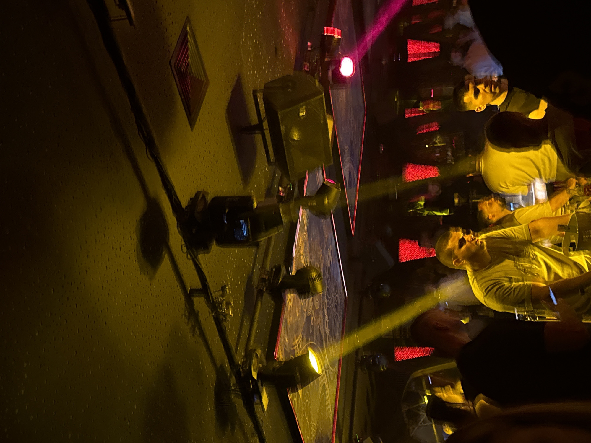 The image depicts an indoor scene, likely a nightclub or bar, with a dark ceiling that has a textured, speckled surface and is illuminated by yellow and pink stage lighting. Several spotlights and speakers are mounted on the ceiling. The lower portion of the image shows several individuals, mostly young men, some of whom are blurred due to motion or low light. They appear to be standing or mingling in a crowded area, with some looking towards the camera or engaged in conversation. The lighting creates dramatic shadows and highlights on their faces and clothing. In the background, there are red-colored lighting fixtures and more people, also mostly blurred. The overall atmosphere is dark and energetic, characteristic of a nightlife venue. The specific location in the city of Sofia, Bulgaria, is not visually discernible from the image.