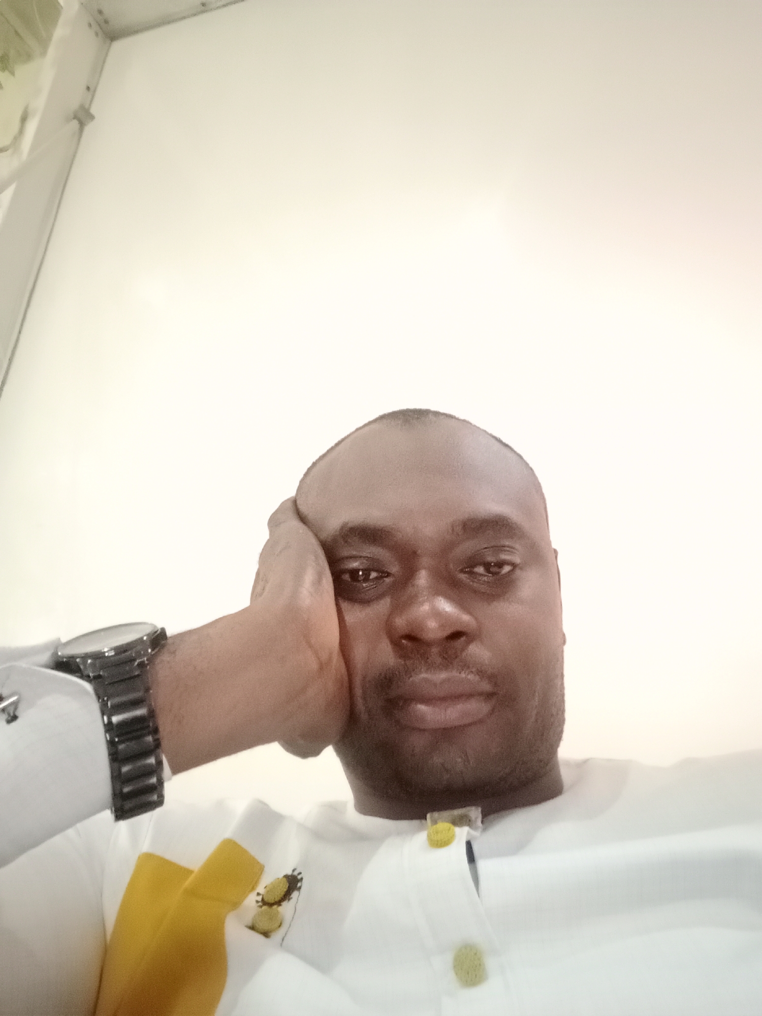 A close-up photograph depicts a dark-skinned man with a shaved head. He is resting his head on his right hand, which has a black wristwatch with a metal band. The man is wearing a white shirt with decorative yellow accents and buttons. The background is a plain, off-white wall. There is no visual information in the photograph to confirm the location as Gwarinpa, Nigeria.