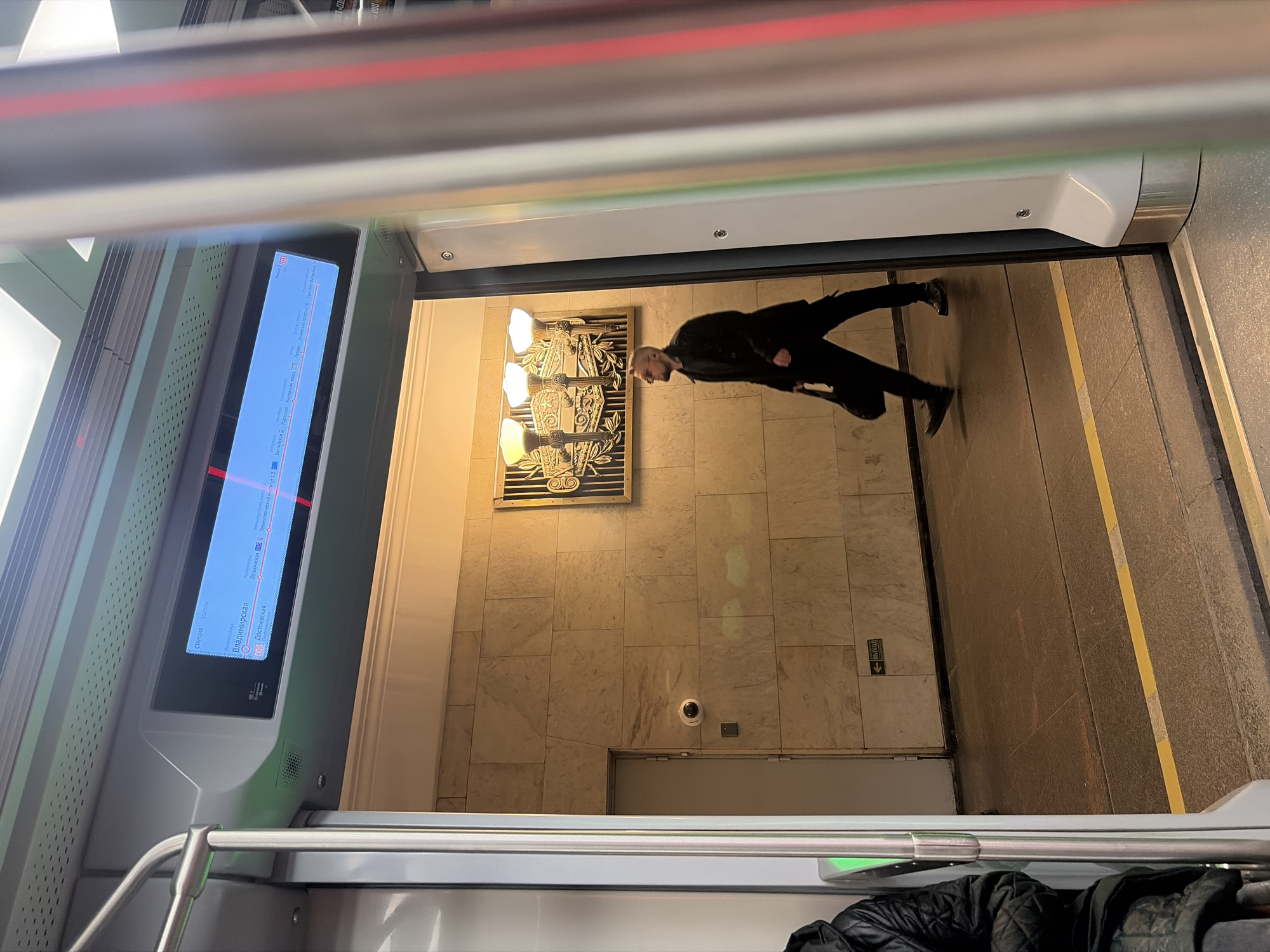 The image is taken from inside a subway car in Saint Petersburg, Russia, showing the doorway opening to a station platform. The camera is likely positioned slightly above the ground.
A man in a dark jacket and trousers is walking away from the subway car onto the platform. On the wall behind him is an artwork and a security camera. The walls are tiled in marble. There are exit signs present.
To the left of the frame, inside the subway car, there is a digital display showing the subway line and the names of upcoming stations including "Vladimirskaya," "Dostoyevskaya," and "Pushkinskaya." Also visible are handrails and the seats of other passengers in the train car. The subway car is modern with red and green accents.
The platform floor has yellow safety lines.