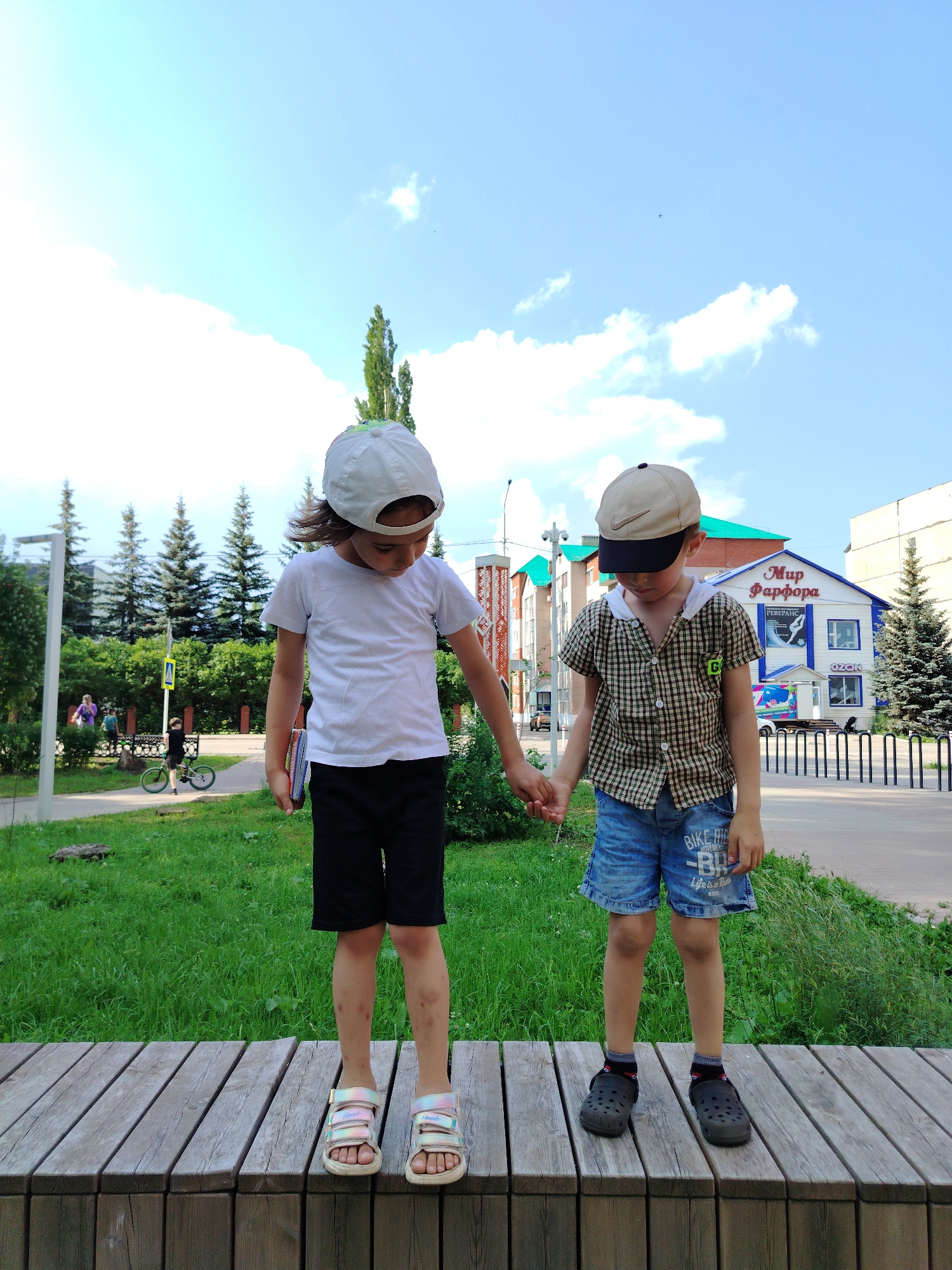Two young children, one wearing a white t-shirt and black shorts, and the other wearing a plaid shirt and denim shorts, stand holding hands on a wooden platform outdoors. The child on the left wears a white cap and colorful sandals, while the child on the right wears a beige and black cap and dark clogs. In the background, a building with the sign "Мир Фарфора" (World of Porcelain) is visible, identifying the location as Yanaul, Russia. The environment includes trees, grass, a sidewalk, and a street with a vehicle and a person on a bicycle in the distance. The sky is blue with scattered clouds.