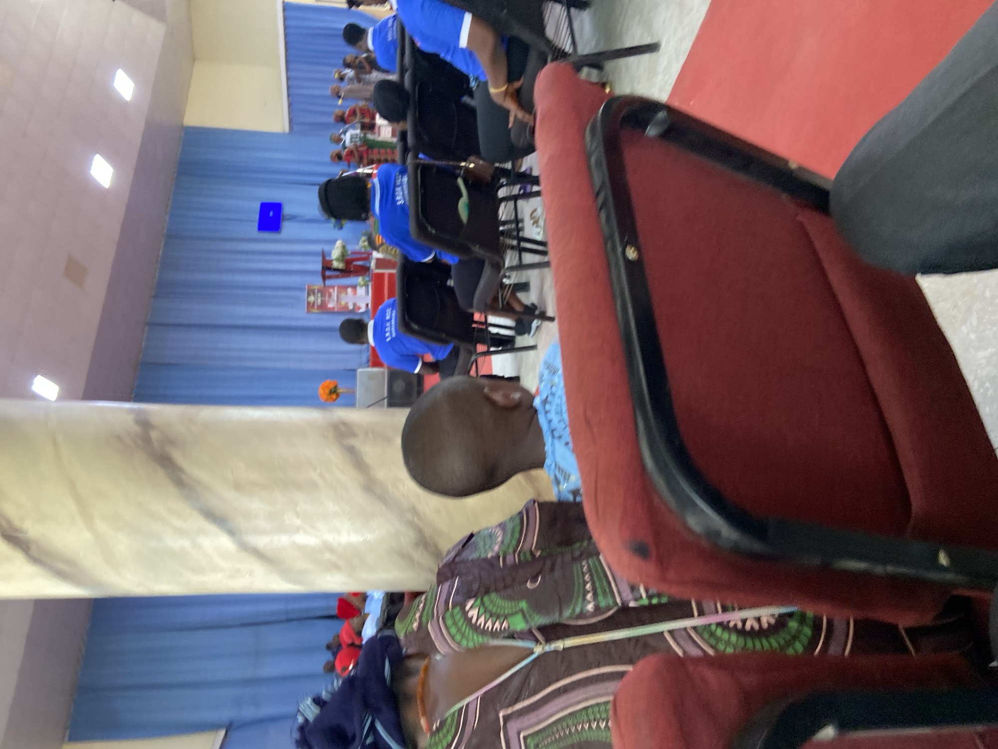 A medium shot taken indoors. Focus is on a seated individual from the back. In front of them, individuals wearing blue shirts are seated in rows. Beyond them, a stage is visible with a blue backdrop and objects. A marbled pillar is also present. The scene appears to be in a church or auditorium setting. It could be Mararaba, Nigeria.