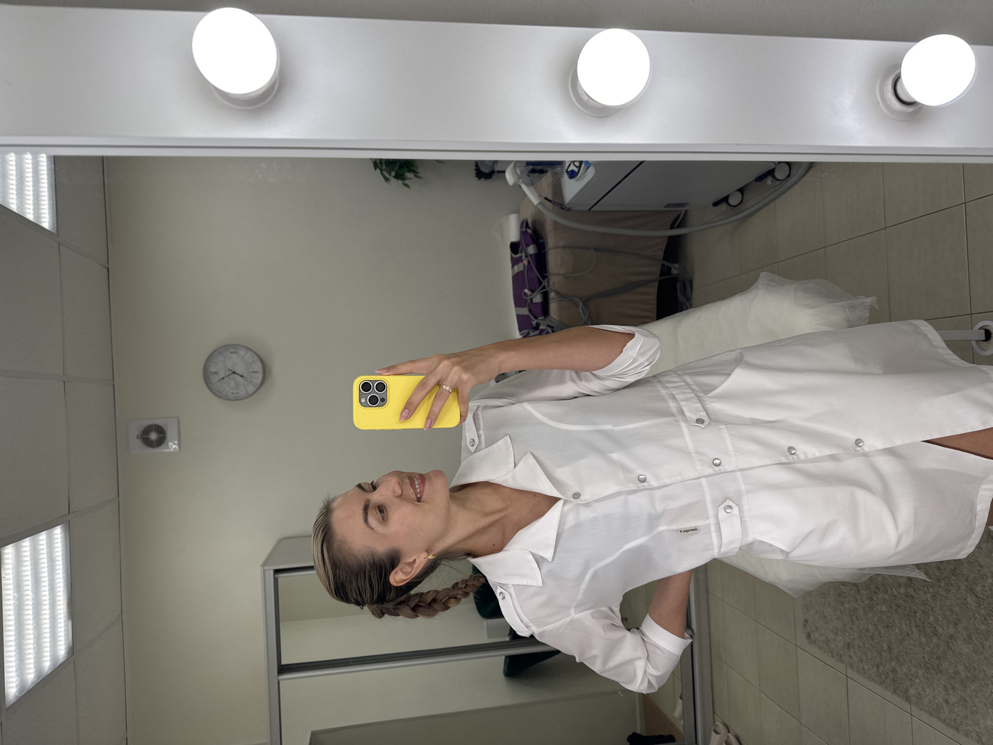 A woman in a white medical or lab coat is captured taking a mirror selfie indoors. She is smiling and looking towards the bright yellow smartphone she holds in her right hand, which features a triple camera setup. Her light-colored hair is styled in a braid, and a ring adorns her right ring finger.
The setting is a professional interior, likely a medical office, clinic, or beauty salon. The selfie is taken in a large mirror framed with several illuminated circular vanity lights along its top edge. Reflected in the mirror are white walls, a white-faced wall clock showing approximately 11:45 or 12:45, and an air vent. The ceiling displays white grid tiles with recessed rectangular fluorescent light panels. Further back in the reflection, pieces of medical or aesthetic equipment are visible, including a treatment bed or chair draped in white linens, and metallic components with hoses or cables. A sliding door wardrobe is also reflected in the background. Part of a textured grey rug or carpet can be seen on the floor.
A small white label on the woman's coat is partially visible, bearing the word "Медицинская," which is Russian for "Medical," confirming the context of her attire.