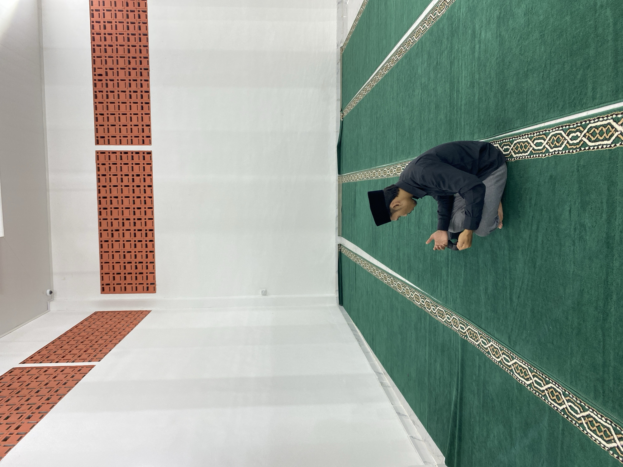 A young man wearing a black peci and a black shirt sits cross-legged on a green carpet with a patterned border in what appears to be a mosque. The room has white walls with decorative brickwork near the ceiling. A security camera is visible on the ceiling. The man is looking down with his hands clasped.