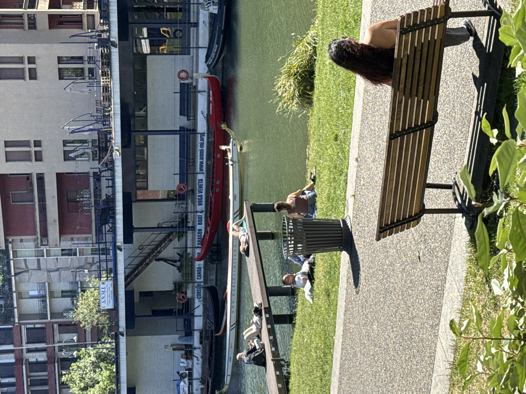 This outdoor shot displays a scene along a canal in Milan, Italy, featuring several people engaging in various activities on a sunny day.
On the right side of the image, a woman sits on a wooden park bench, with her back to the camera. The bench is situated on a paved area next to a patch of green grass.
Adjacent to the paved area is a canal with several boats docked along a wooden pier. People are seen lounging and interacting on the pier. On the right, a man is resting with his arm extended over a small trash bin. Further to the left, another man in a white shirt lies face up. Two more individuals are sitting towards the left, and someone wearing a black jacket is visible at the end of the pier.
Docked along the pier are several boats, including a traditional gondola and dragon boats with signage in Italian. The building behind the pier is multi-storied and features a sign indicating that it is the A.N.M.I.L. B association, offering courses in canoeing, dragon boat, and rowing. The building's design incorporates both residential and commercial elements.