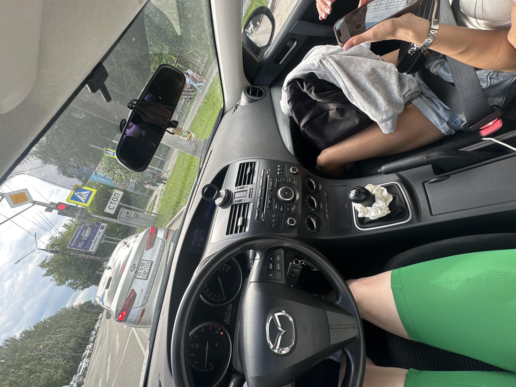 The image is taken from inside a car, looking out the windshield. The car is stopped at a traffic light in the city of Perm, Russia. In front of the car is a silver Kia with a Russian license plate. Traffic is lined up on the road, and pedestrians can be seen on the sidewalk. The interior of the car is visible, including the steering wheel, dashboard, and gear shift. A person's legs, wearing green shorts, are visible in the driver's seat. Another person, in the passenger seat, is holding a phone. The sky is partly cloudy.