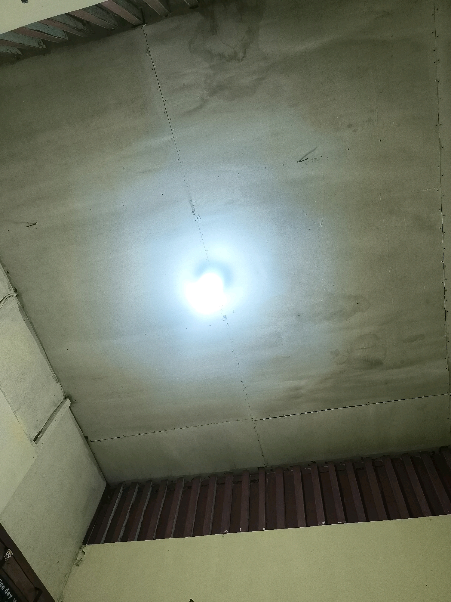 This is a ceiling in what appears to be a room under construction or undergoing renovation. The ceiling is made of large, light-colored panels, likely drywall or a similar material, joined together by visible seams and secured with numerous small nails or screws. There are some signs of wear and tear, including water stains and an uneven texture in places. A bright light source, possibly a bare bulb, is illuminating the center of the ceiling, creating a strong glare and obscuring some of the finer details in its vicinity. The edges of the ceiling meet walls that are painted a pale yellow or cream color. In the lower part of the image, there is a visible gap between the ceiling and the wall, revealing wooden beams or structural elements that are dark reddish-brown. The overall impression is one of an unfinished or utilitarian space. The location is Timika Jaya, Indonesia.