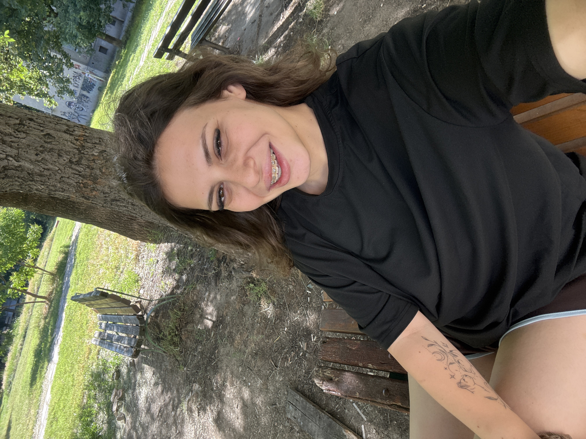 A young woman with brown hair, braces, and large eyelashes is sitting on a wooden park bench in Plovdiv, Bulgaria. She is wearing a black t-shirt and shorts. Her left arm is visible, showing a tattoo of a cat with celestial elements. The background features a tree, grass, other park benches, and a building with graffiti. The lighting suggests it is daytime and sunny.