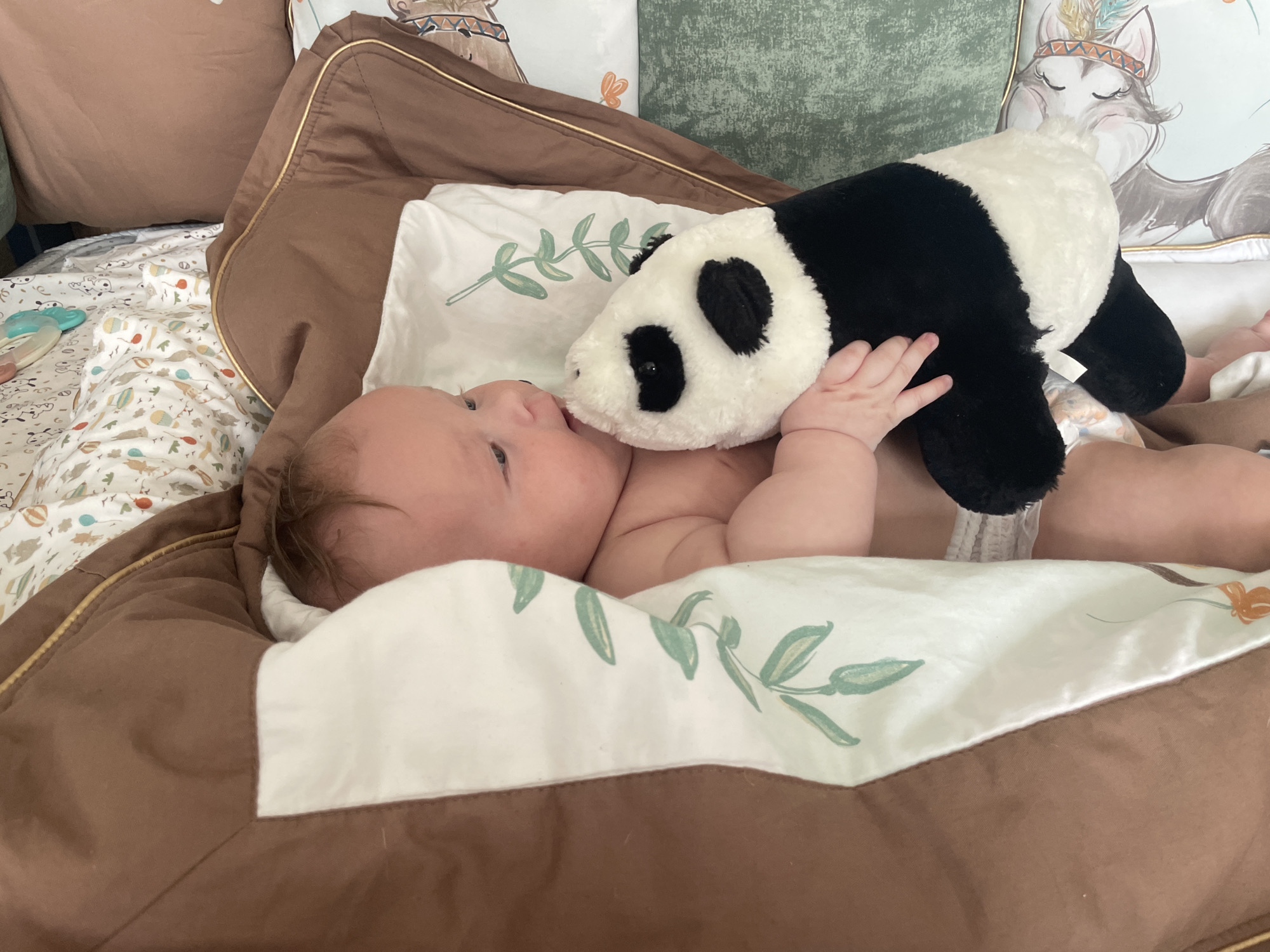 A baby is lying on a brown blanket, propped up by pillows, and playing with a stuffed panda. The baby is wearing a diaper and has a pacifier in its mouth. The stuffed panda is black and white and is being held by the baby's hand. The background is a blur of pillows and blankets. The image appears to be taken indoors in a bedroom. It is not possible to determine the location in the city of Stavropol, Russia from the image.