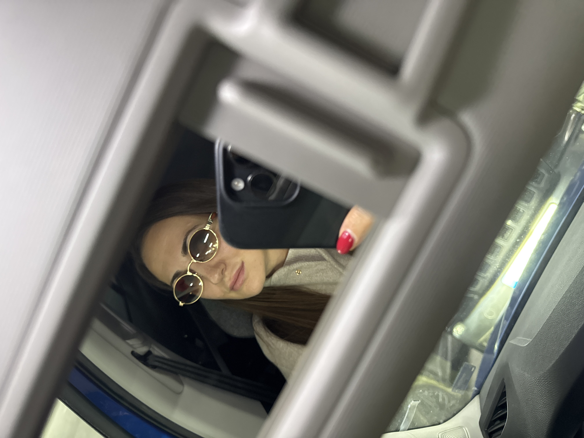 A woman wearing round sunglasses is reflected in a car visor mirror. She holds a black smartphone, visible by its camera lenses, with her right hand, where her fingernail is painted red. The interior of the car is seen, including parts of the dashboard and the side of the door. The car is likely in a location where there is artificial lighting, as suggested by the reflections. The location of the city of Varna, Bulgaria is not evident from the image.