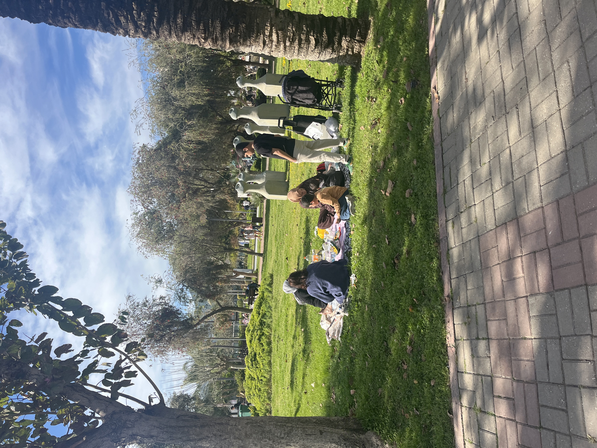 The image depicts a park scene in Alanya, Turkey. Several individuals are picnicking on the green grass. A man stands near a blanket, while two other people are sitting on the ground. Adjacent to them are several statues. The backdrop includes trees and a partially visible blue sky with clouds. A paved walkway runs along the edge of the green area.