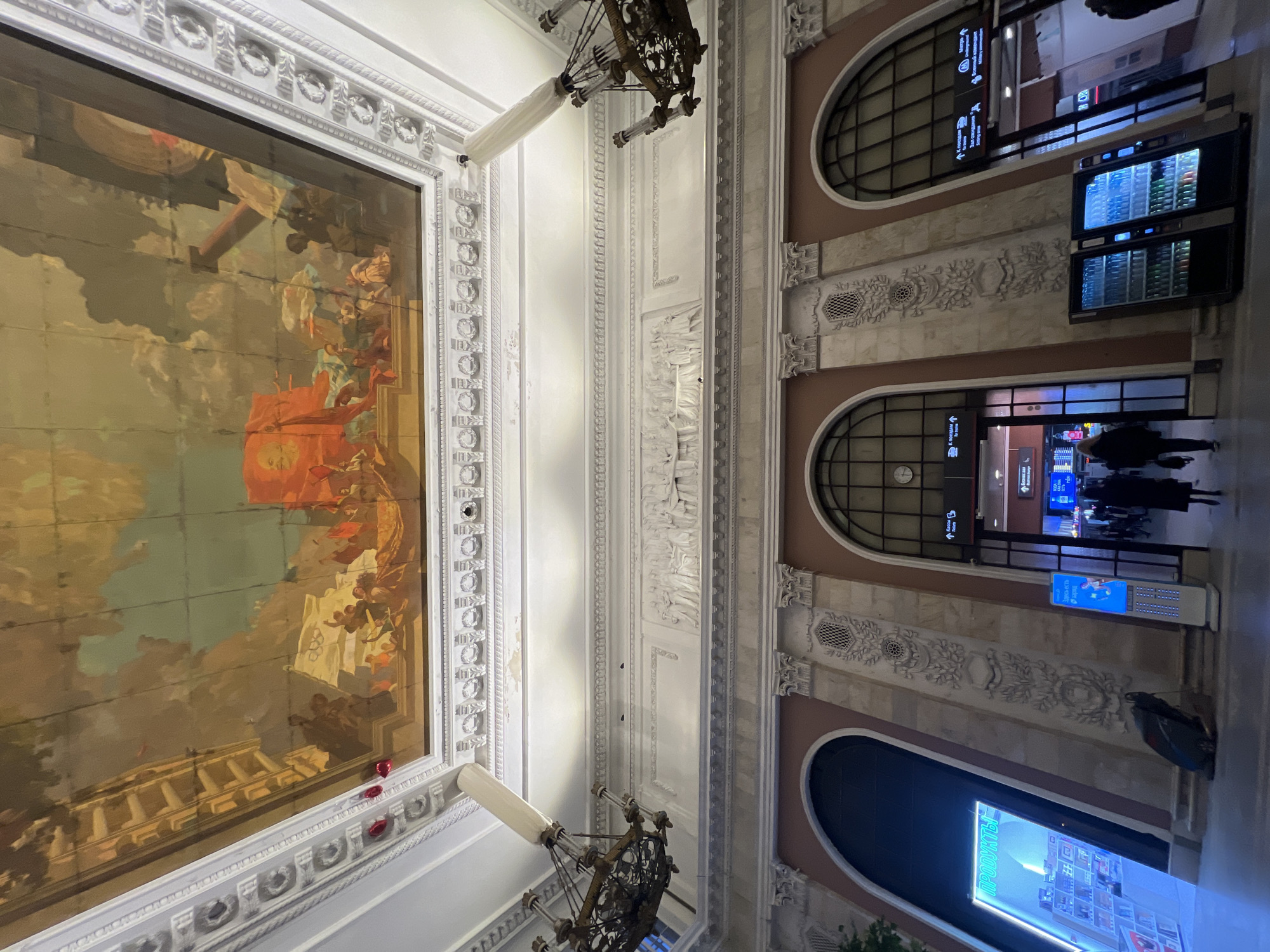 The image shows the interior of a railway station in Saint Petersburg, Russia. A large, ornate painting decorates the ceiling, featuring classical figures and a prominent red flag. The architectural details include arched windows, decorative moldings, and vintage-style chandeliers. Digital signage and vending machines indicate a modern transportation hub. People are seen walking through the station, suggesting transit activity.