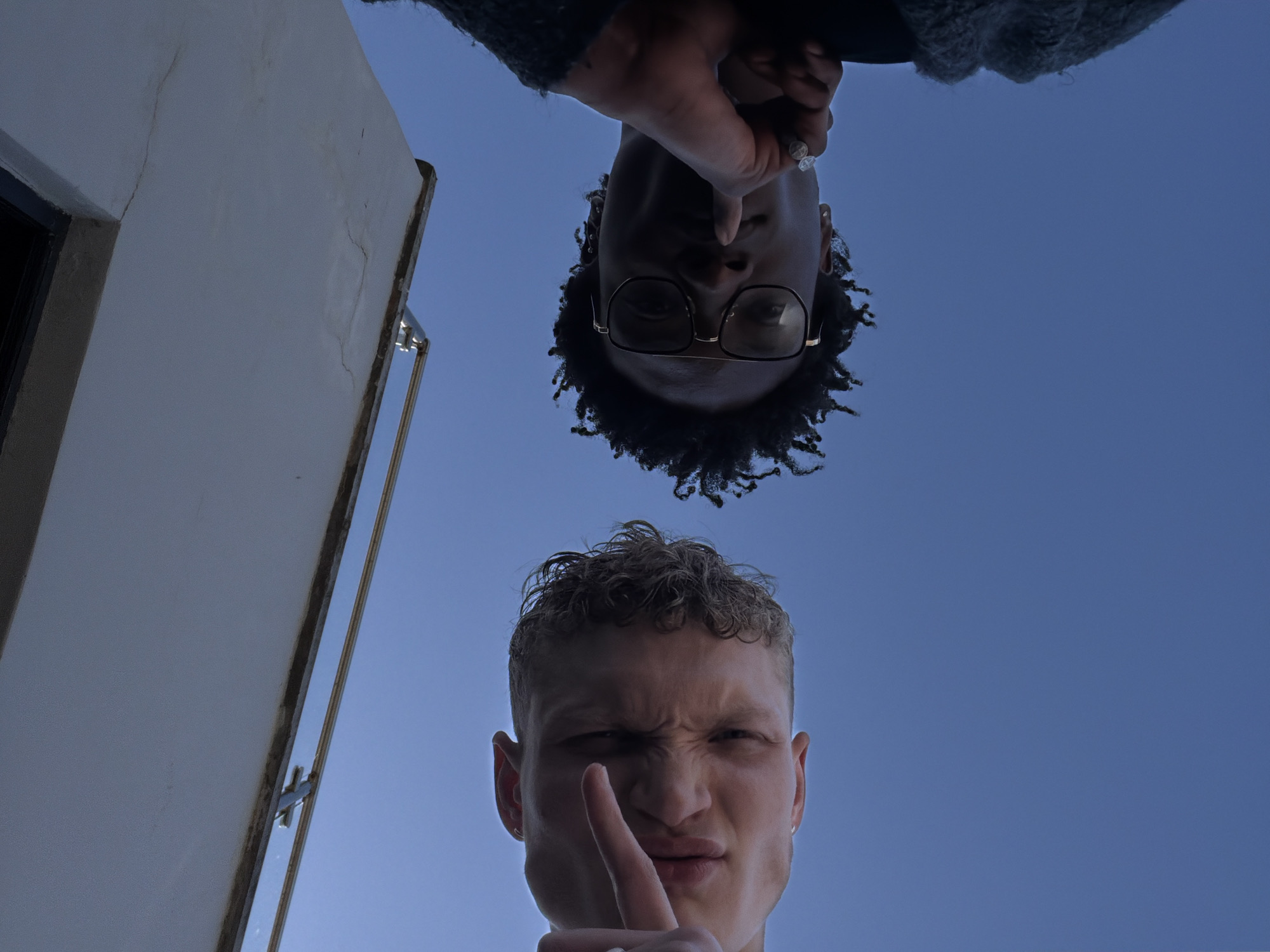 The image presents a striking low-angle, worm's-eye view, looking straight up into a clear blue sky. Two men are captured in this unusual perspective, along with parts of a building structure.
At the top of the frame, appearing inverted, is a man with dark skin, dark curly hair, and eyeglasses. His hands are clasped near his chin, partly obscuring his face. He is wearing what appears to be a dark, textured garment.
Below him, positioned in the lower half of the frame and oriented conventionally, is another man with light skin and light, curly hair. He is making a "shushing" gesture, with his right index finger raised to his lips. His mouth is slightly puckered, and his eyes are squinted, conveying a playful, secretive, or mischievous expression.
The background is a vibrant, cloudless blue sky, indicating a bright daytime setting. On the left side of the frame, a segment of a plain white building wall or structure is visible, extending vertically upwards. On the right, a metallic railing or support structure is partially seen, further defining a built environment. While the context suggests London, United Kingdom, there are no specific visual cues within the image to confirm this location. No text is visible in the media file.
