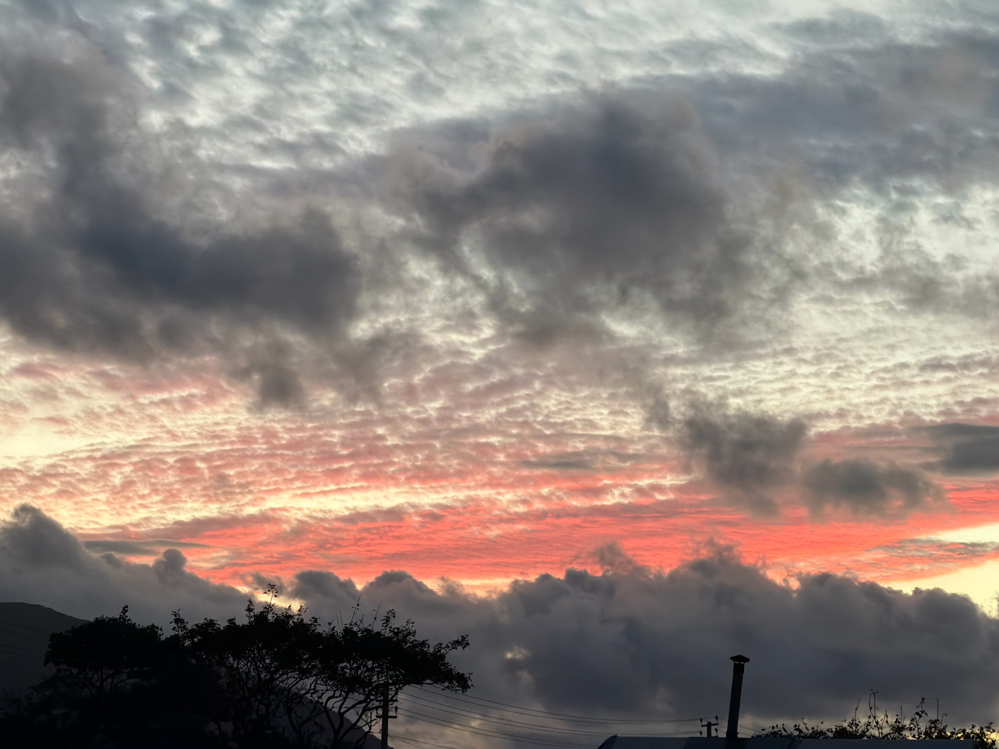 The image captures a dramatic sunset over Yuzhno-Kurilsk, Russia, with a sky filled with a complex arrangement of clouds. The lower portion of the sky is ablaze with vibrant coral and pink hues, illuminated by the setting sun. Above this, darker, heavier clouds in shades of gray and charcoal obscure parts of the horizon, creating a sense of depth and contrast. Higher still, wispy, lighter clouds streak across the sky, tinged with pale gray and white, reflecting the fading sunlight. In the foreground, the silhouettes of trees and vegetation are visible, along with the dark outlines of what appear to be rooftops and a chimney, indicating an inhabited area. The overall impression is one of a powerful natural display of light and shadow over a semi-urban or rural landscape.