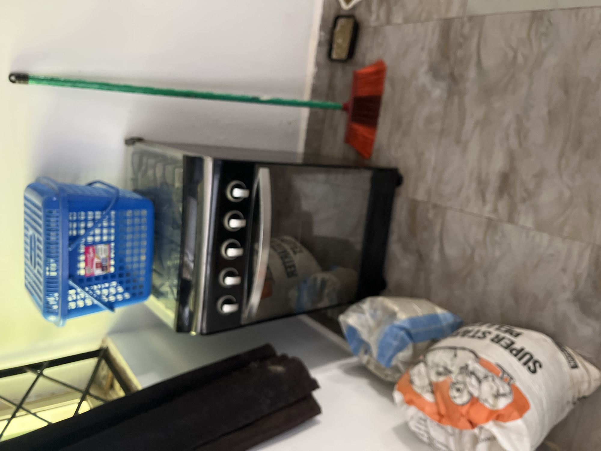 The image shows a corner of a room with a black gas stove. On top of the stove, there is a blue plastic basket. To the right of the stove, leaning against the wall, is a green broom with red bristles. In front of the stove, on the floor, are two large bags, one with an orange and white label that reads "SUPER STAR PET". The floor appears to be tiled with a marble-like pattern. To the left of the stove, a dark curtain hangs down. The overall scene suggests a domestic or storage area, possibly in a residential building in Jos, Nigeria.
