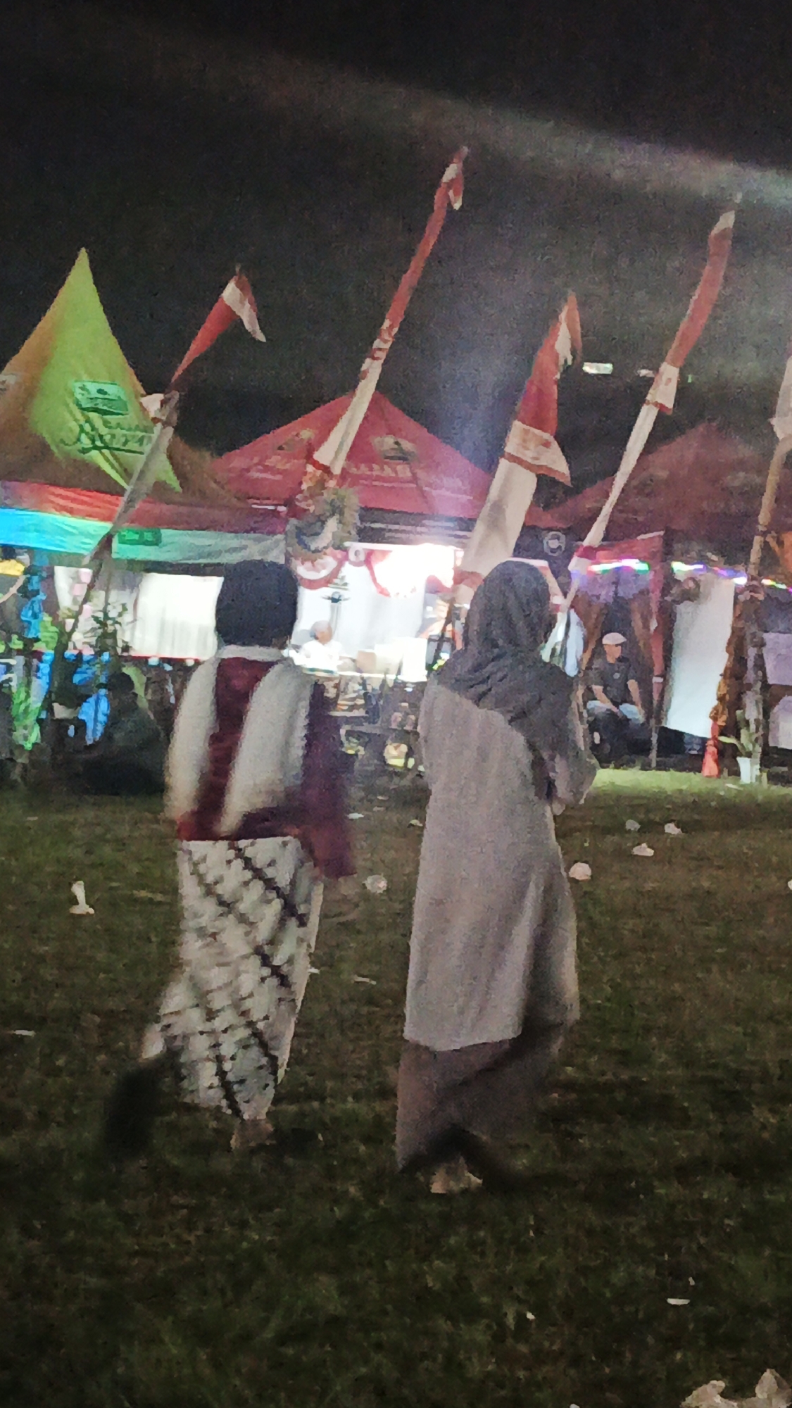 Two individuals, one wearing a white and maroon patterned garment and the other a grey hijab and dress, walk across a grassy area at night. Behind them, several tents are visible, some adorned with flags featuring red and white stripes and Arabic script. The scene suggests a nighttime event or festival, possibly in Ketemasdungus, Indonesia. The lighting is artificial, casting shadows on the ground. The overall atmosphere is dimly lit, characteristic of an outdoor evening gathering.